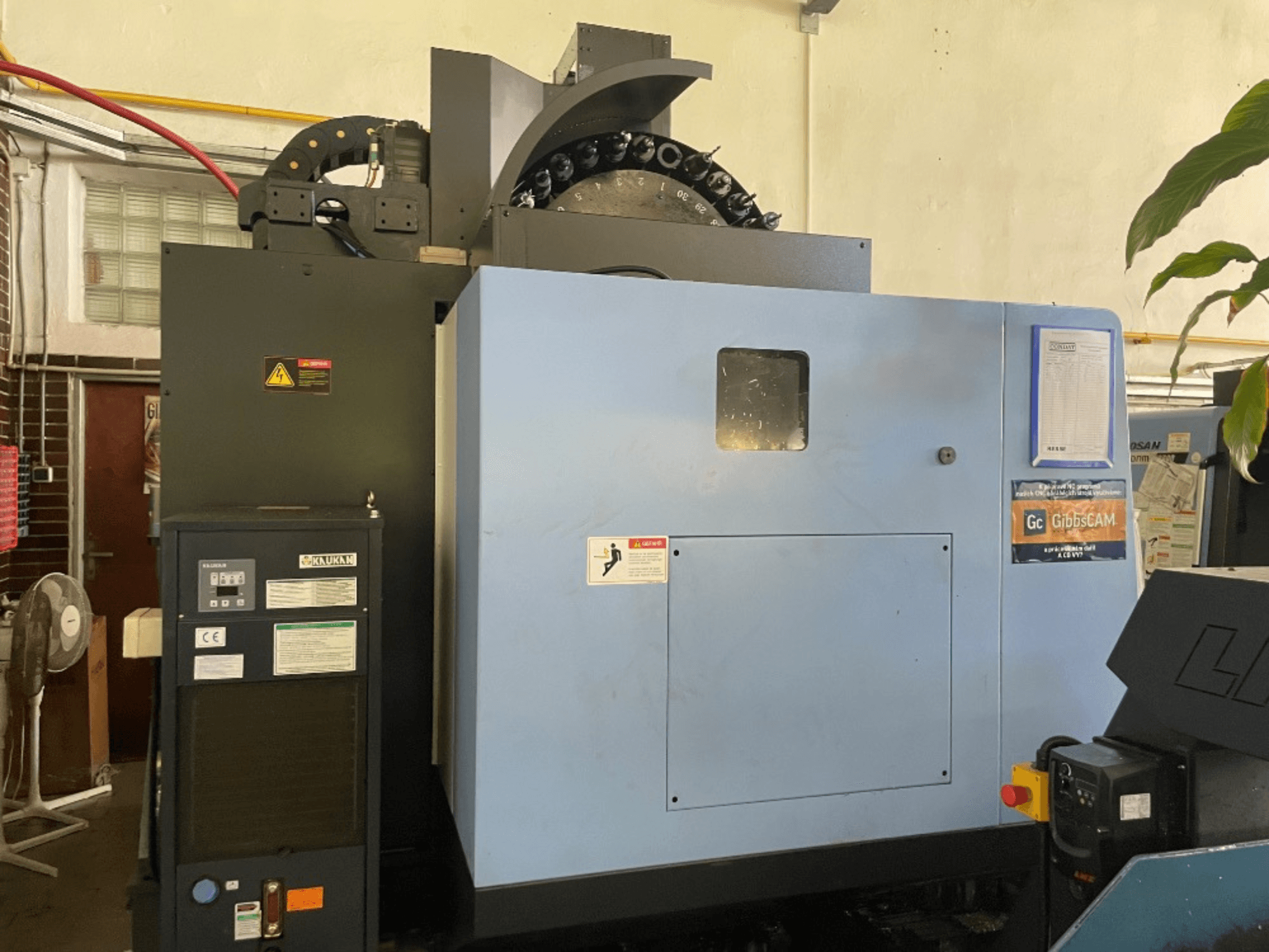 Frontvy av en DOOSAN DNM 5700 vertikal fleroperationsmaskin, med kontrollpanel, säkerhetsetiketter och maskinfunktioner.