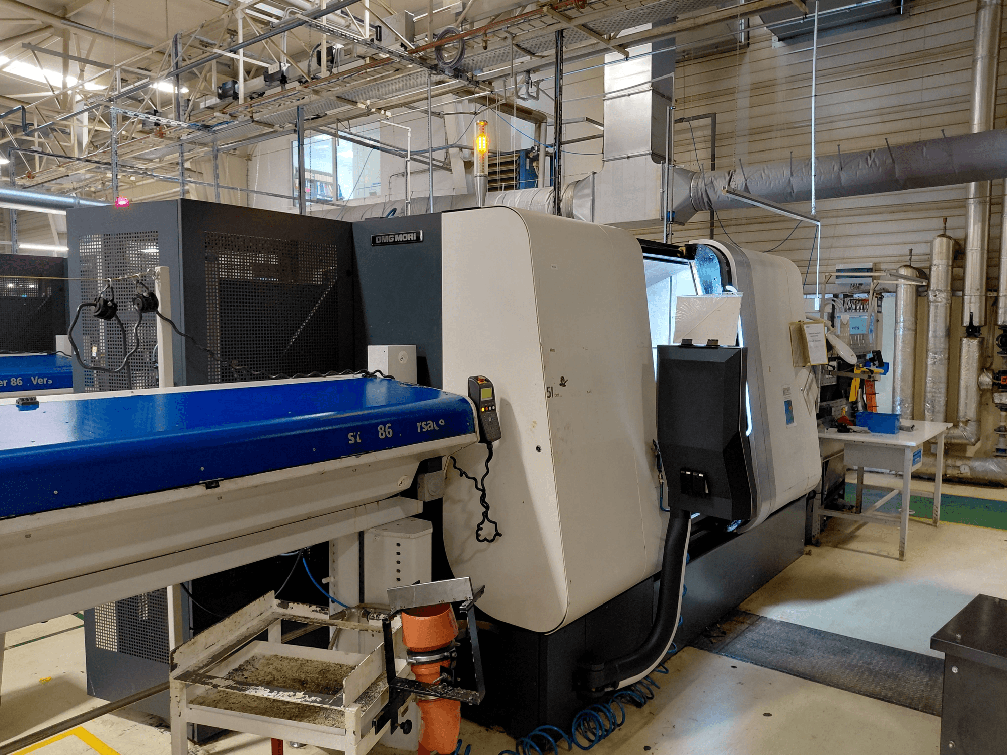 DMG MORI Sprint 65 linjär flerspindlig svarv, sidovy med kontrollpanel och transportör synlig i en verkstadsmiljö.
