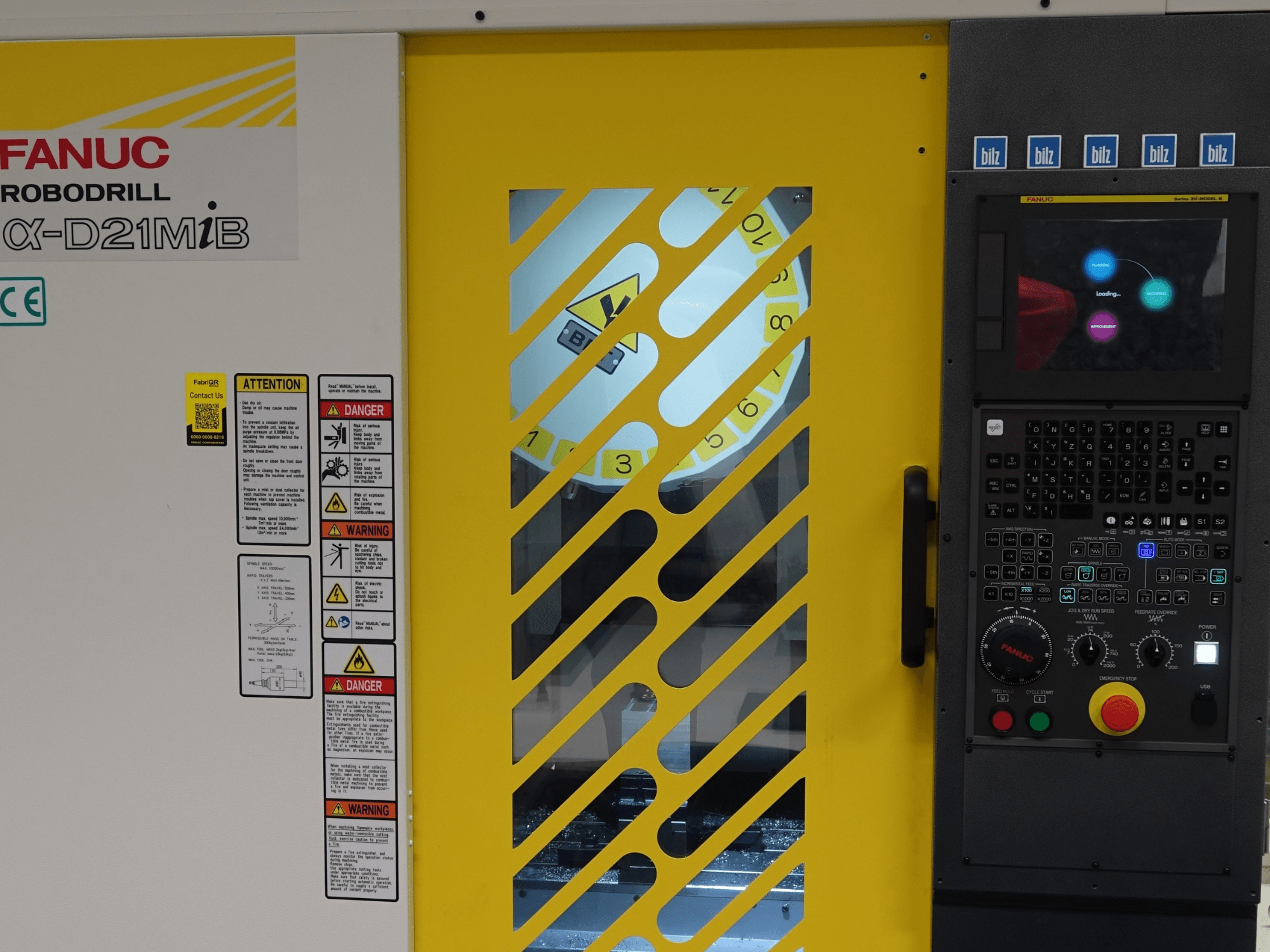 FANUC α-D21MiB vertikalt bearbetningscenter sett framifrån, med säkerhetsvarningar, kontrollpanel och gul säkerhetsdörr.