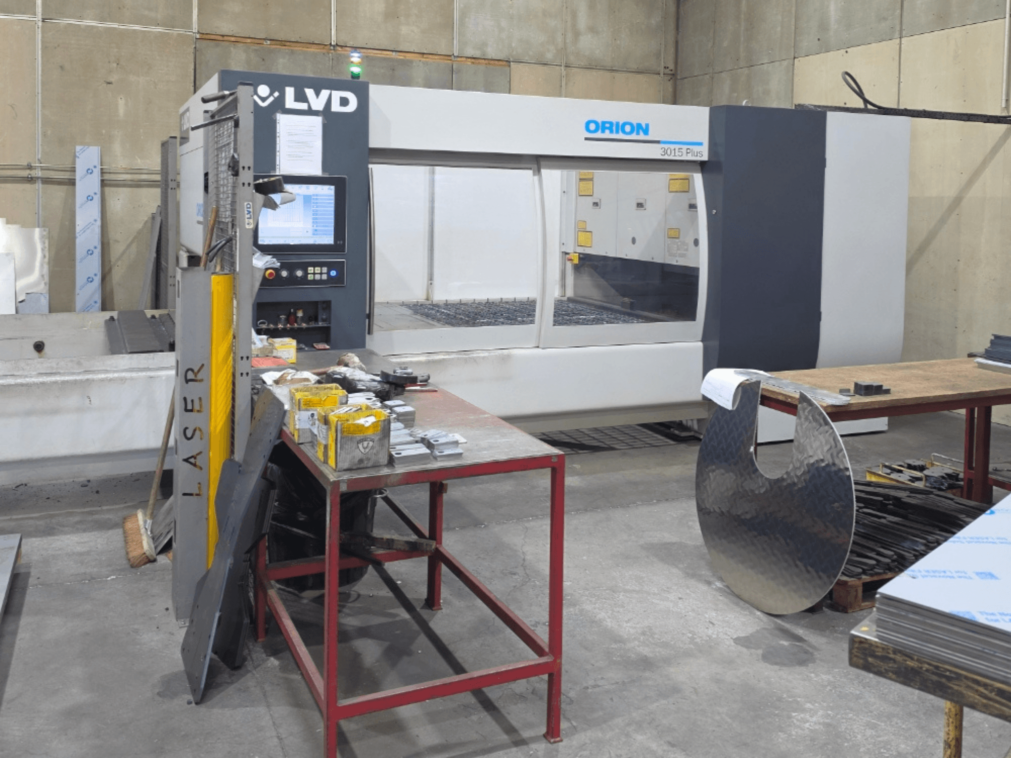 LVD Orion 3015 Plus CO₂-laserskärmaskin, framifrån med kontrollpanelen; omgiven av metalldelar på ett arbetsbord.
