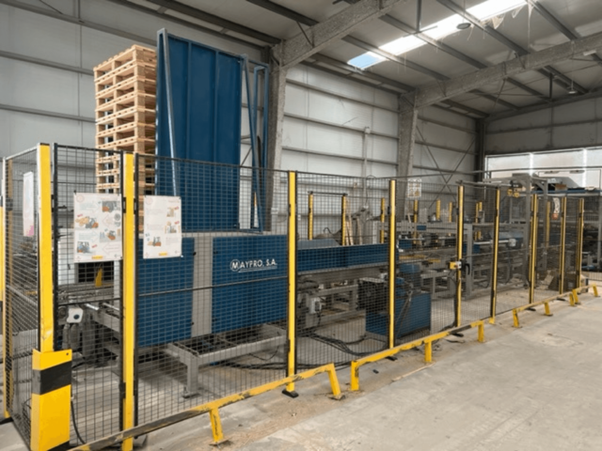 Industriell pallpackningsmaskin från Maypro S.A. i en lagerlokal, med skyddsnät och manöverreglage, vy från sidan.