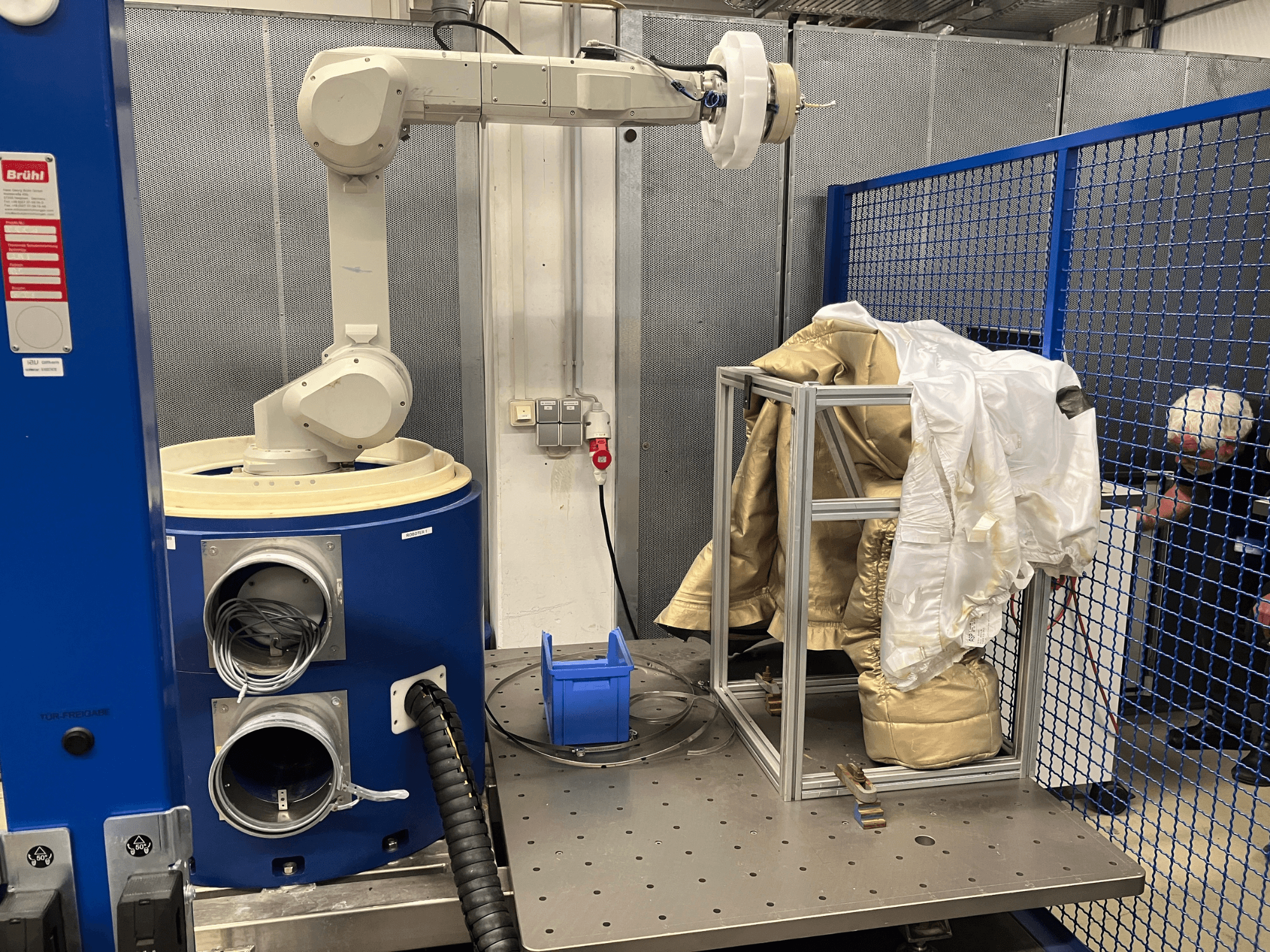 Industriell robotarm bredvid blå cylindrisk maskin som håller tygplagg på en metallram, vy från sidan.