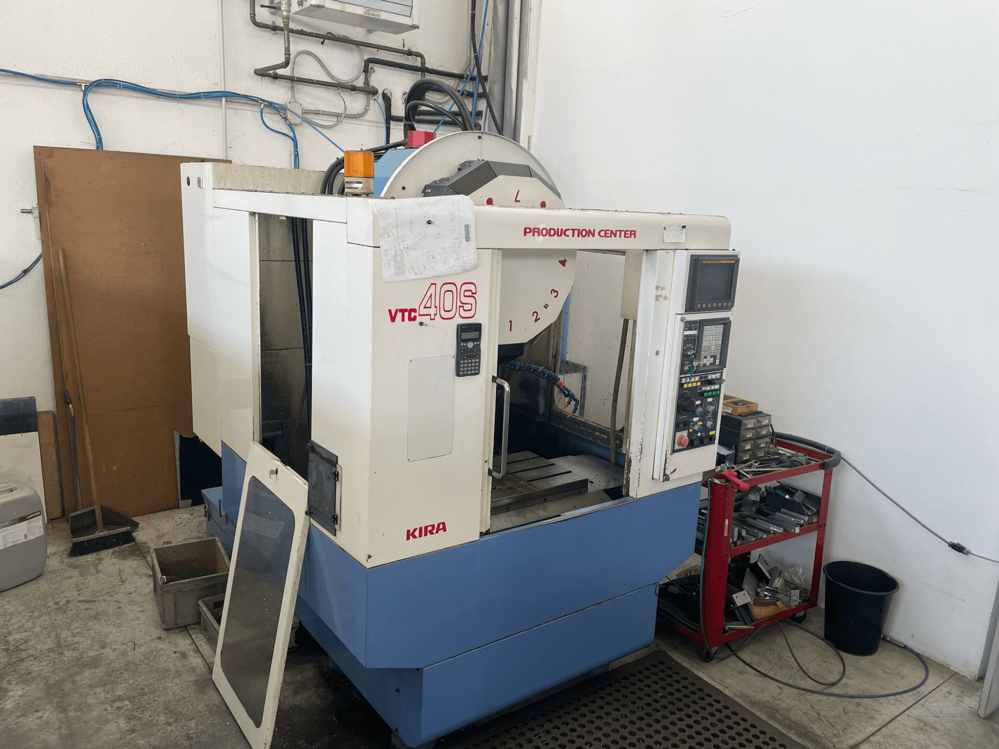 Bearbetningscenter KIRA VTC 40S i sidovy, med kontrollpanel, öppen dörr och verktyg på en förvaringsvagn i närheten.