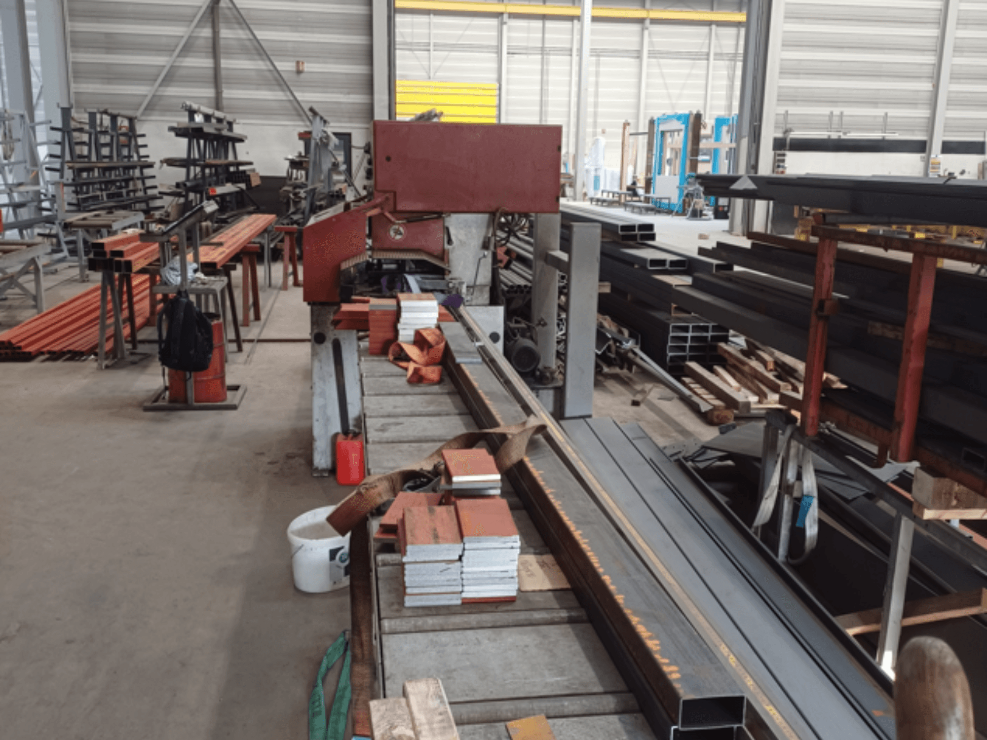 Tillverkningsverkstad med olika metallprofiler, med en röd sågmaskin i förgrunden och staplade material.