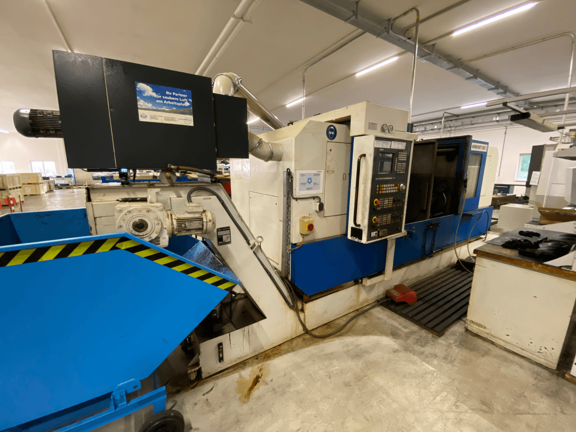 Monforts DNC 5 svarv-fräscenter, sidovy som visar kontrollpanelen, det blå spånfacket och detaljer på maskinen i en verkstadsmiljö.