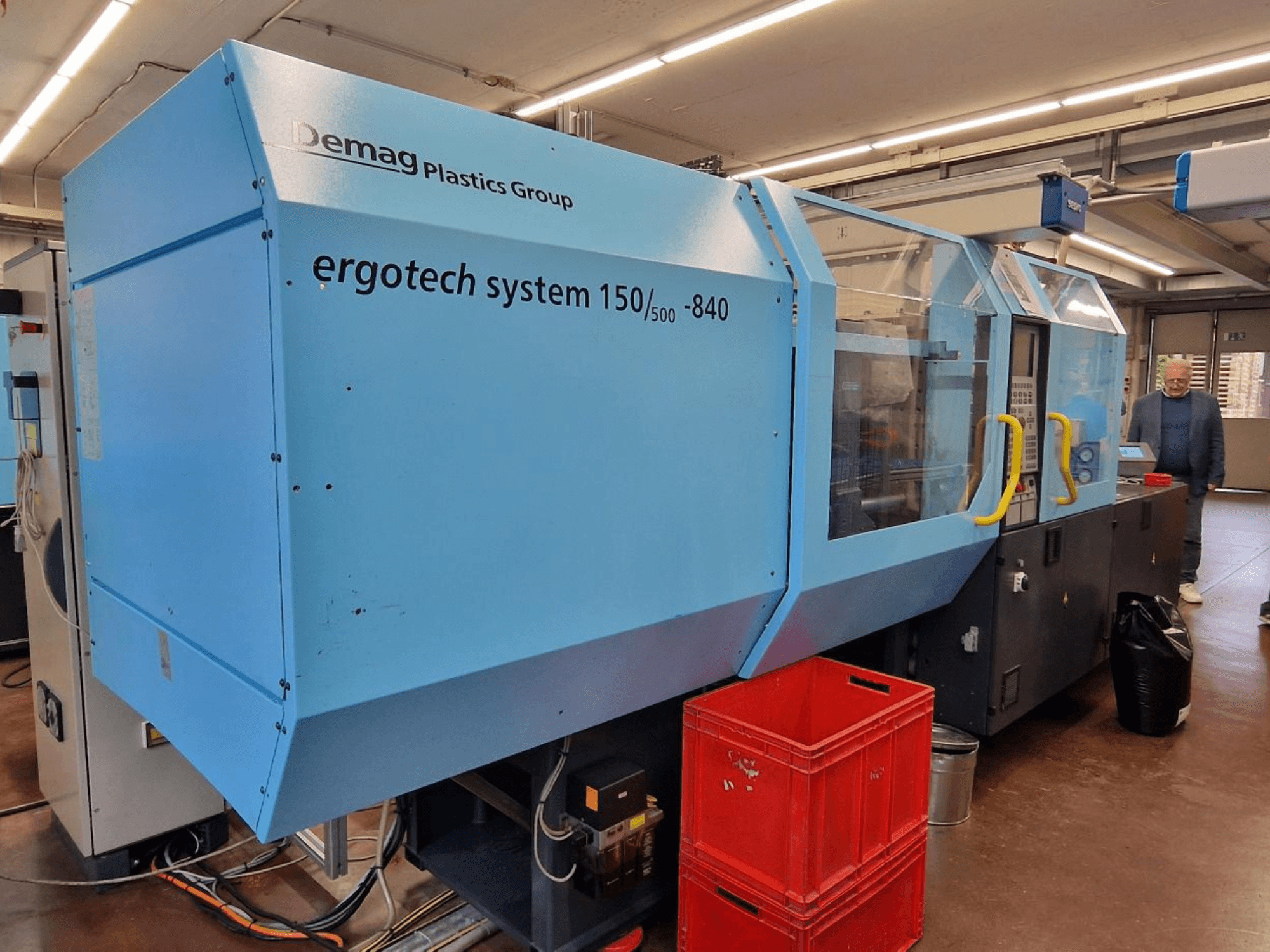 Demag Ergotech System 150/500-840 formsprutningsmaskin i blått, vy från sidan, med lådor och operatör i bakgrunden.