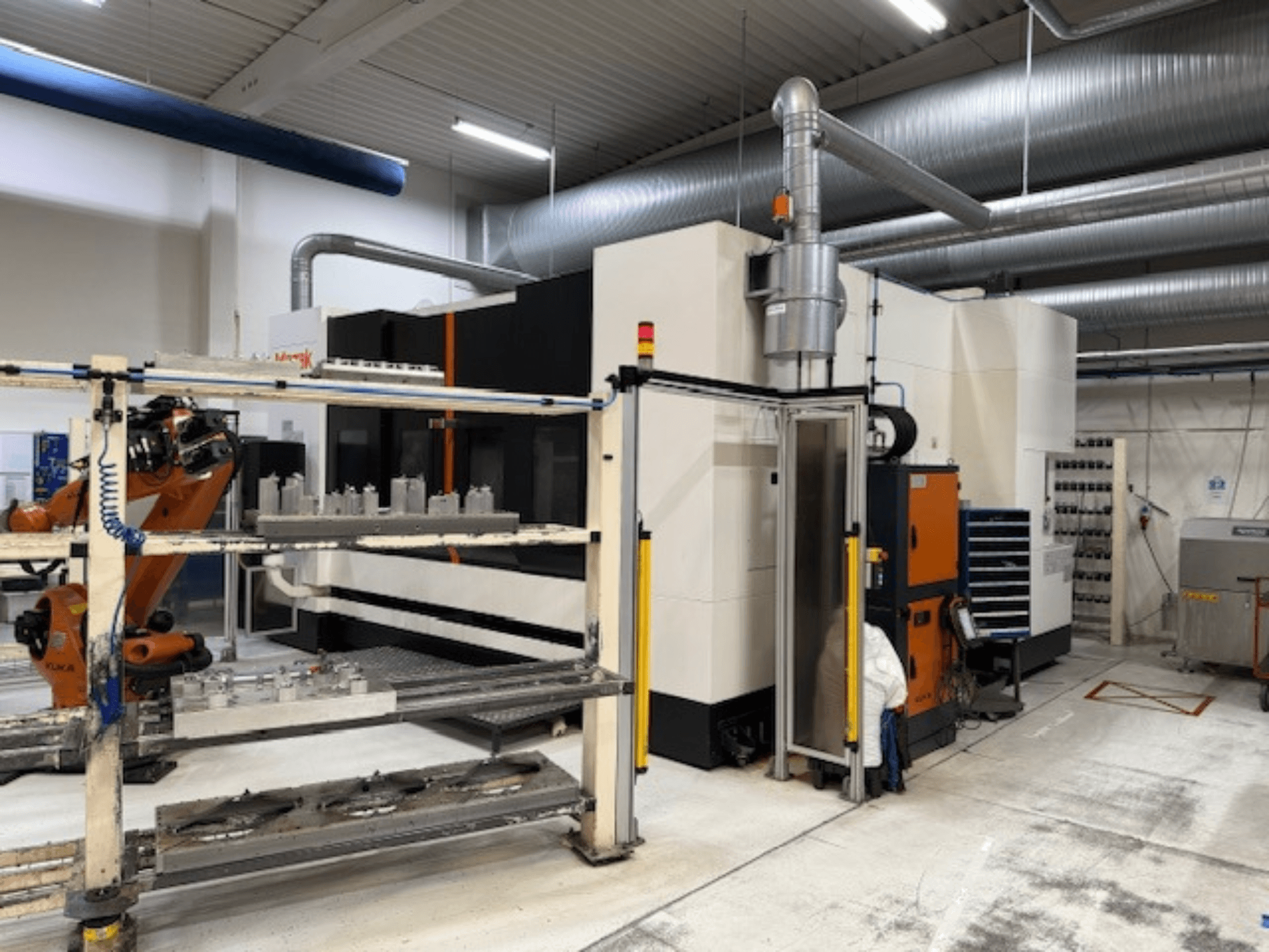 Industriell CNC-maskin med robotarm och säkerhetsbarriärer, med en frontvy av installationen i ett luftigt arbetsutrymme.