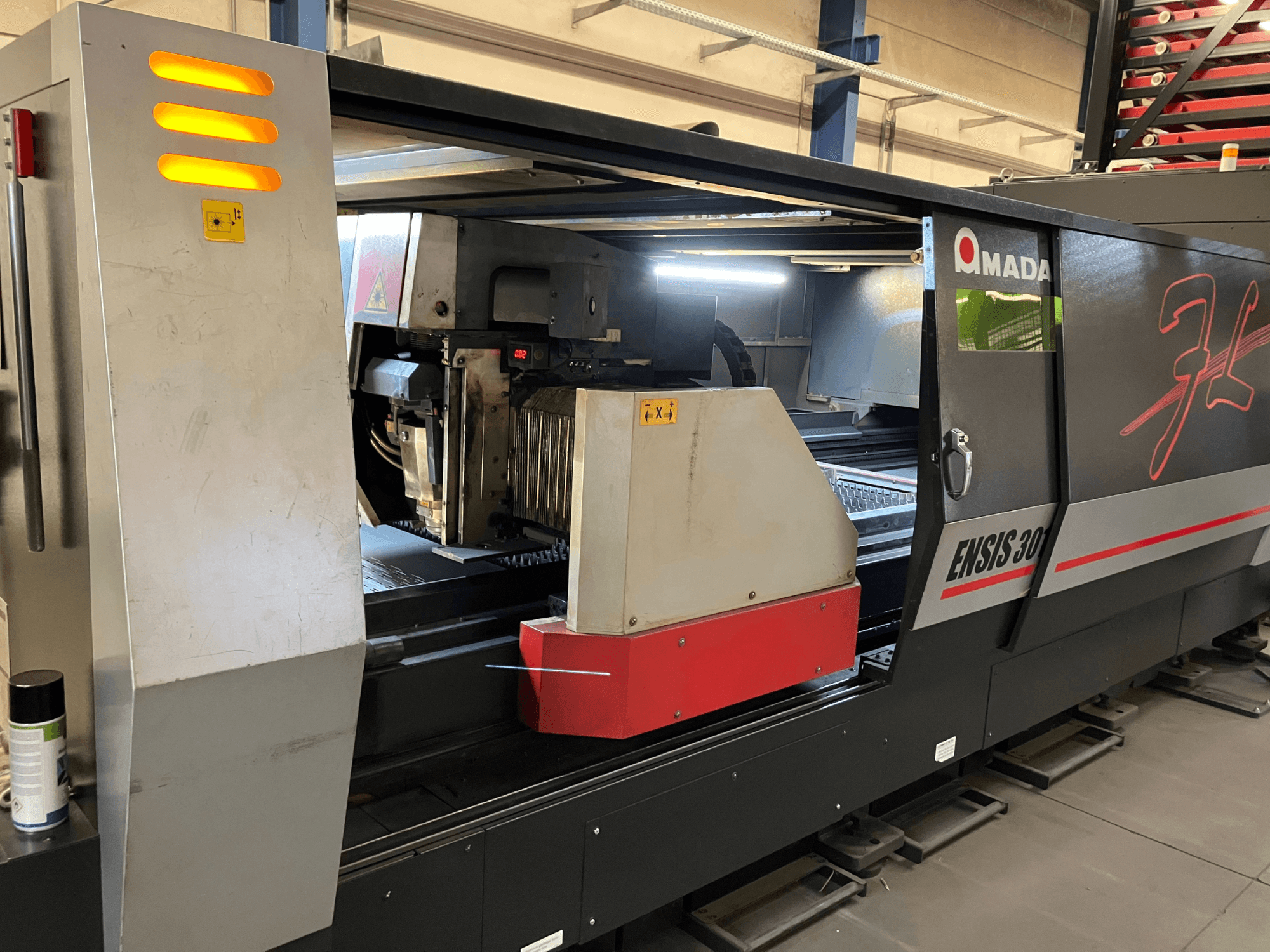 AMADA ENSIS 3015 AJ fiberlaserskärmaskin, framifrån, visar skärhuvud och manöverreglage inuti en metallram.