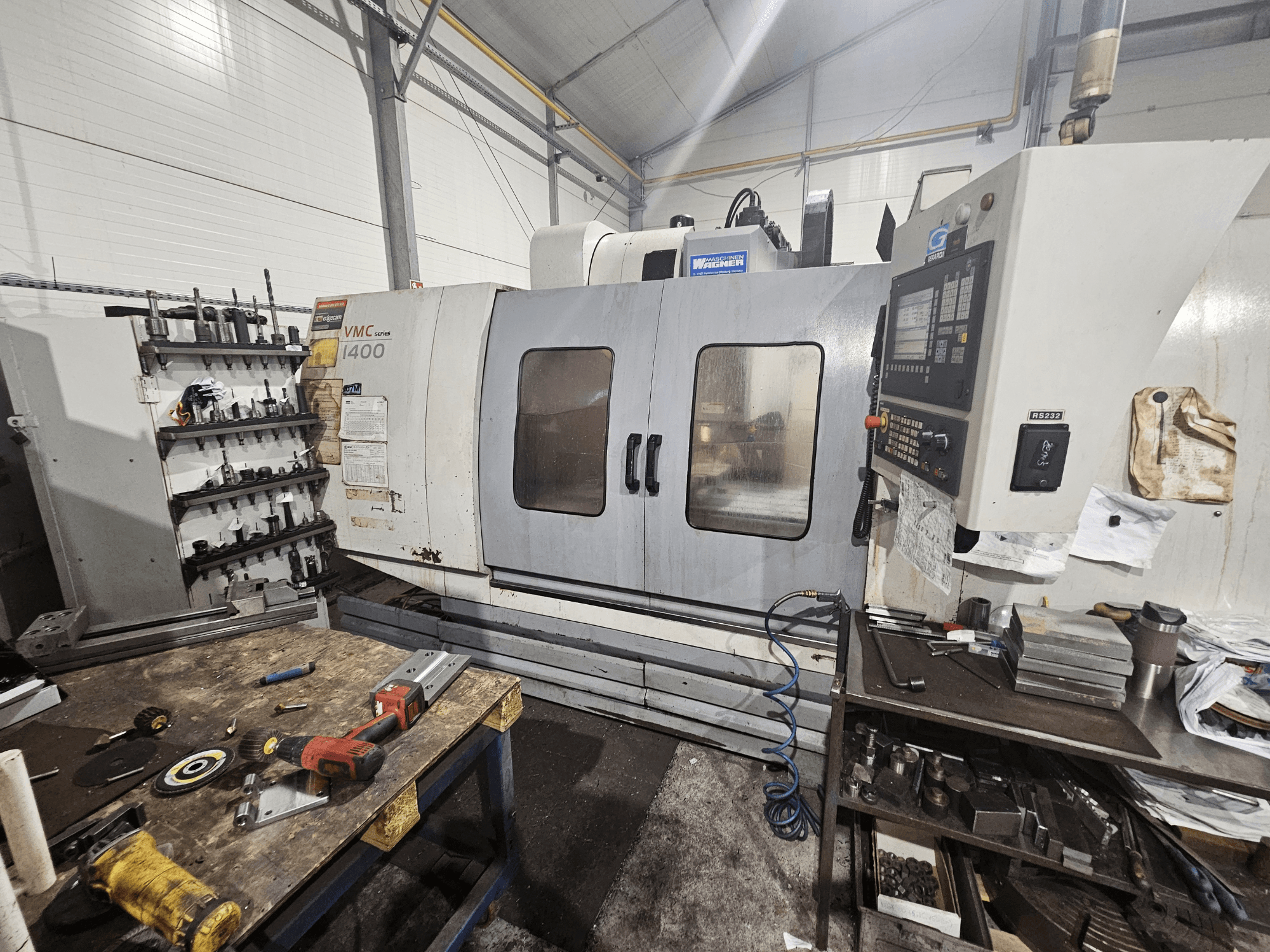 VMC 1400 CNC-fräsmaskin i en verkstad, vy framifrån; verktyg och material utspridda på arbetsbänken i närheten.
