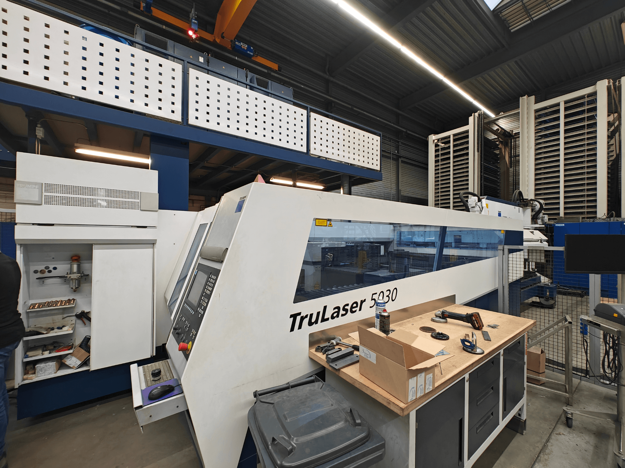 TruLaser 5030-maskin i en verkstad, framifrån med synlig kontrollpanel, omgiven av verktyg och utrustning.