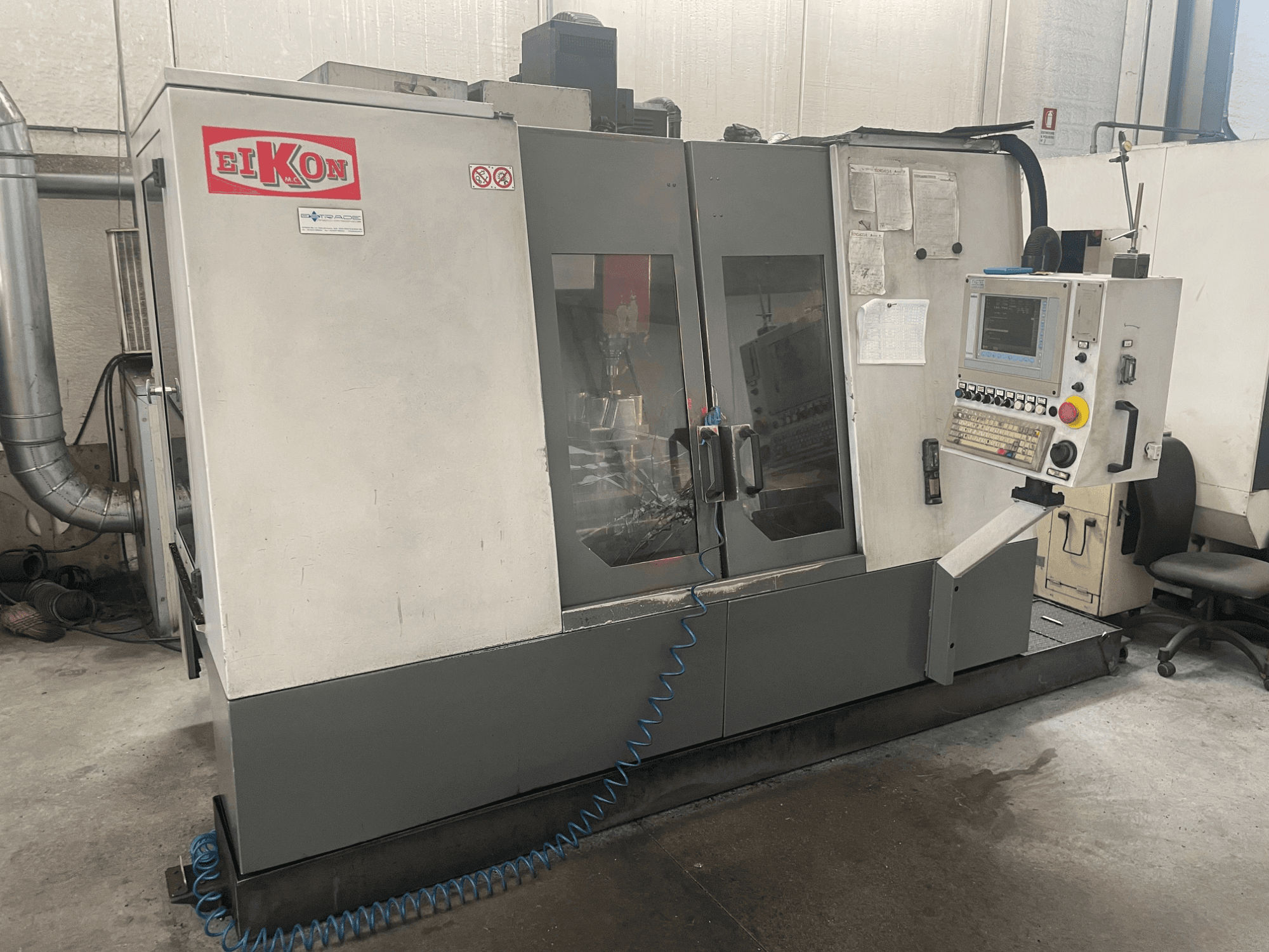 CNC-maskin från EIKON sedd framifrån med kontrollpanel, stora glasdörrar och en blå luftslang på golvet.