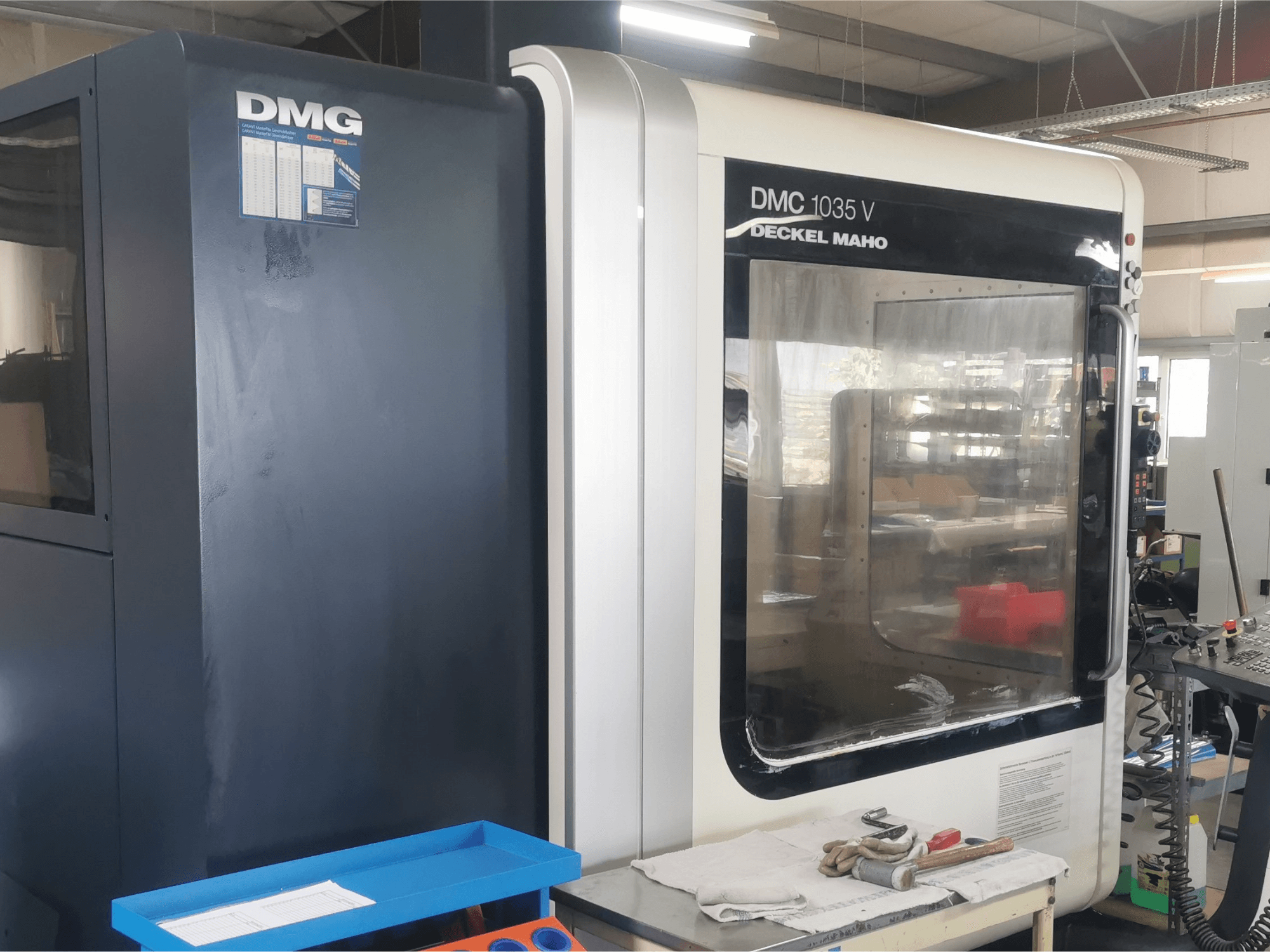 DMG DMC 1035 V fräsmaskin i en verkstad, framifrån med blå verktygslåda och handskar på ytan.