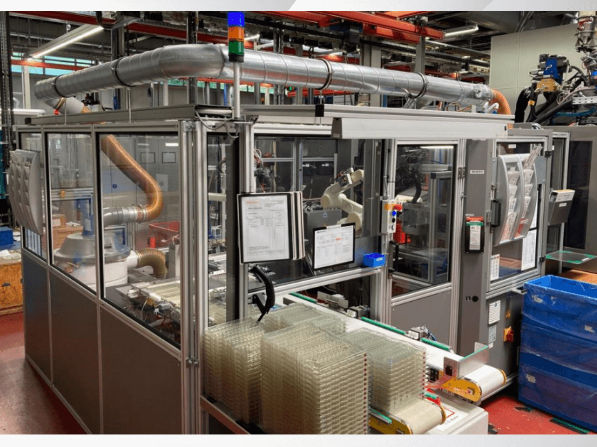 Automatiserad monteringsmaskin med robotarm, genomskinliga sidopaneler och staplade behållare med klara föremål, framifrån.