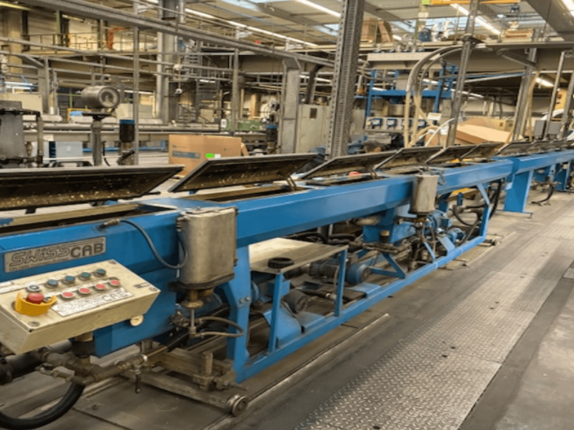Blå SwissCab industrimaskin i fabriksmiljö, med manöverknappar och ett transportsystem, vy från sidan.