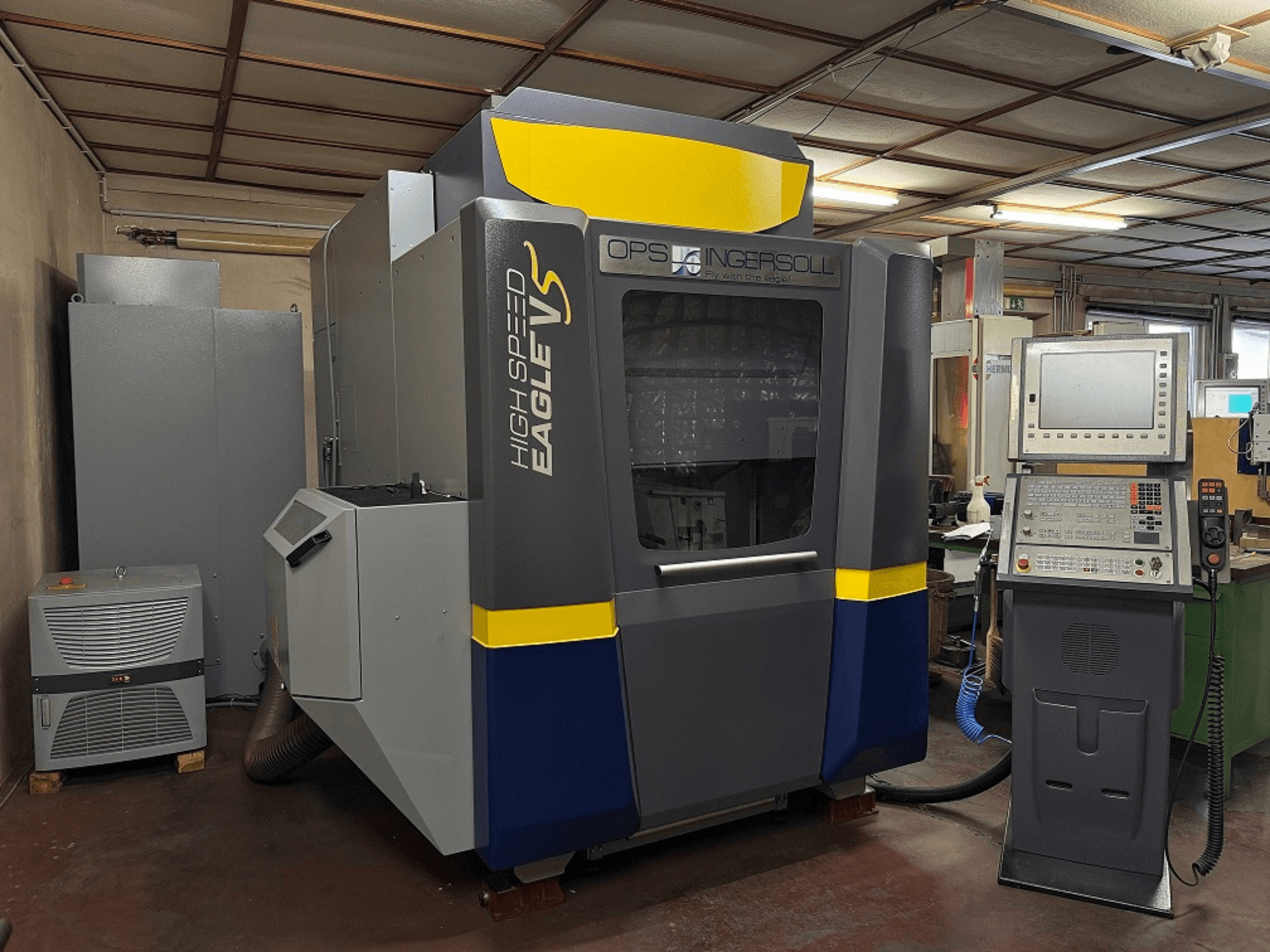 High Speed Eagle V-maskin från OPS Ingersoll, vy från sidan, med kontrollpanel och intilliggande utrustning i industriell miljö.