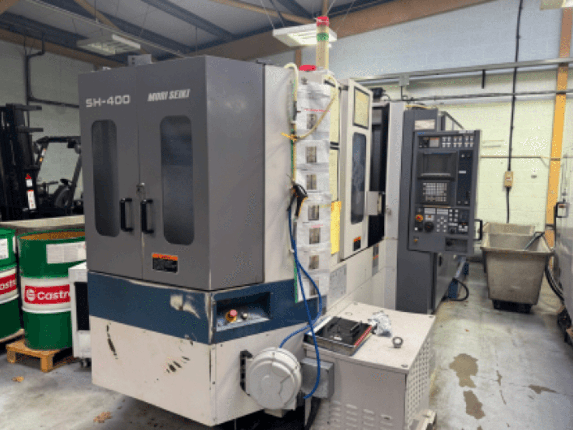 Moni Seiki SH-400 CNC-maskin, sidovy som visar kontrollpanel, dörrar och omgivande industrimiljö.