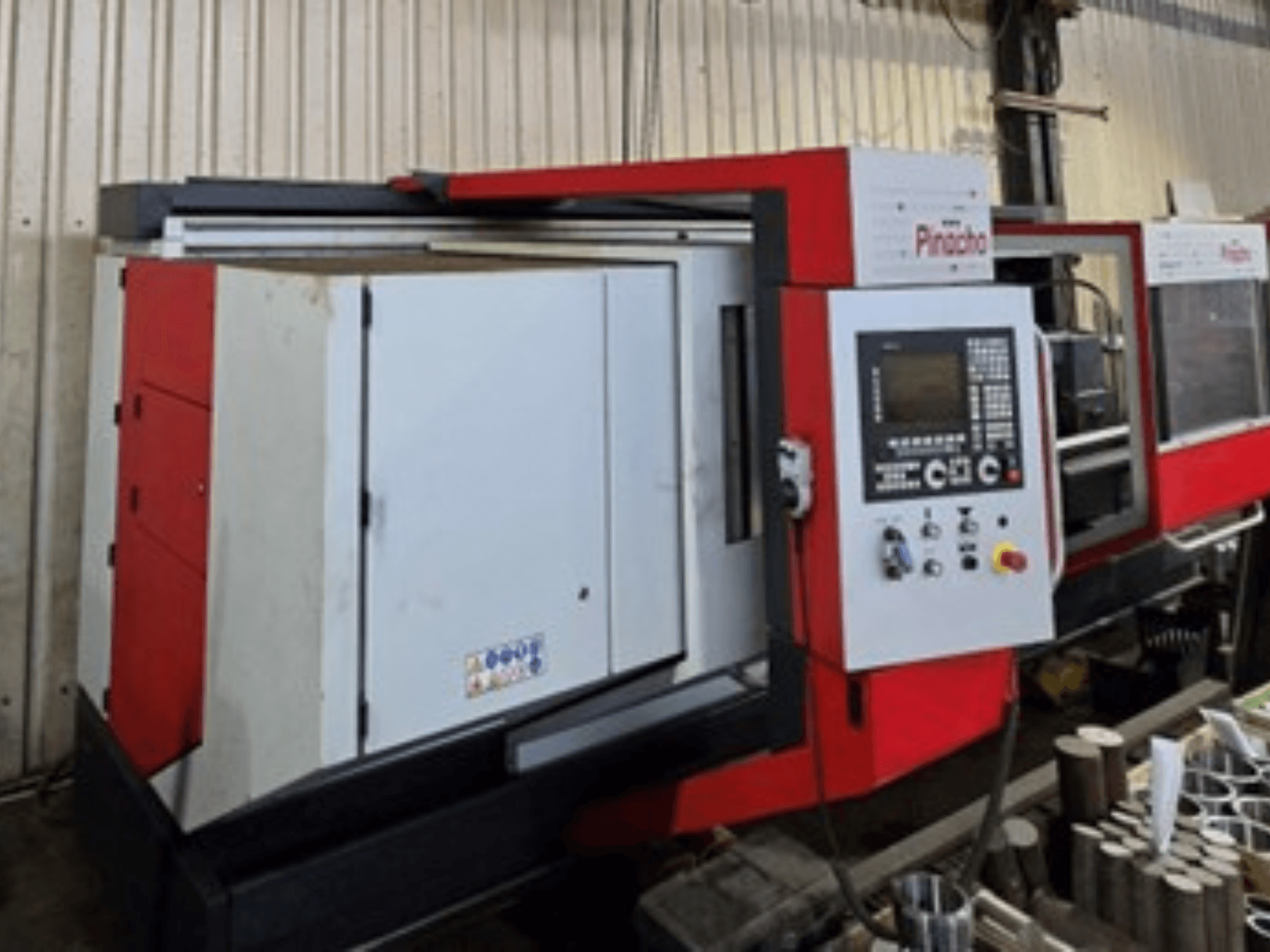 Pinacho CNC-maskin i rött och vitt, vy från sidan, med kontrollpanel och material på golvet, som visar driftsuppställning.