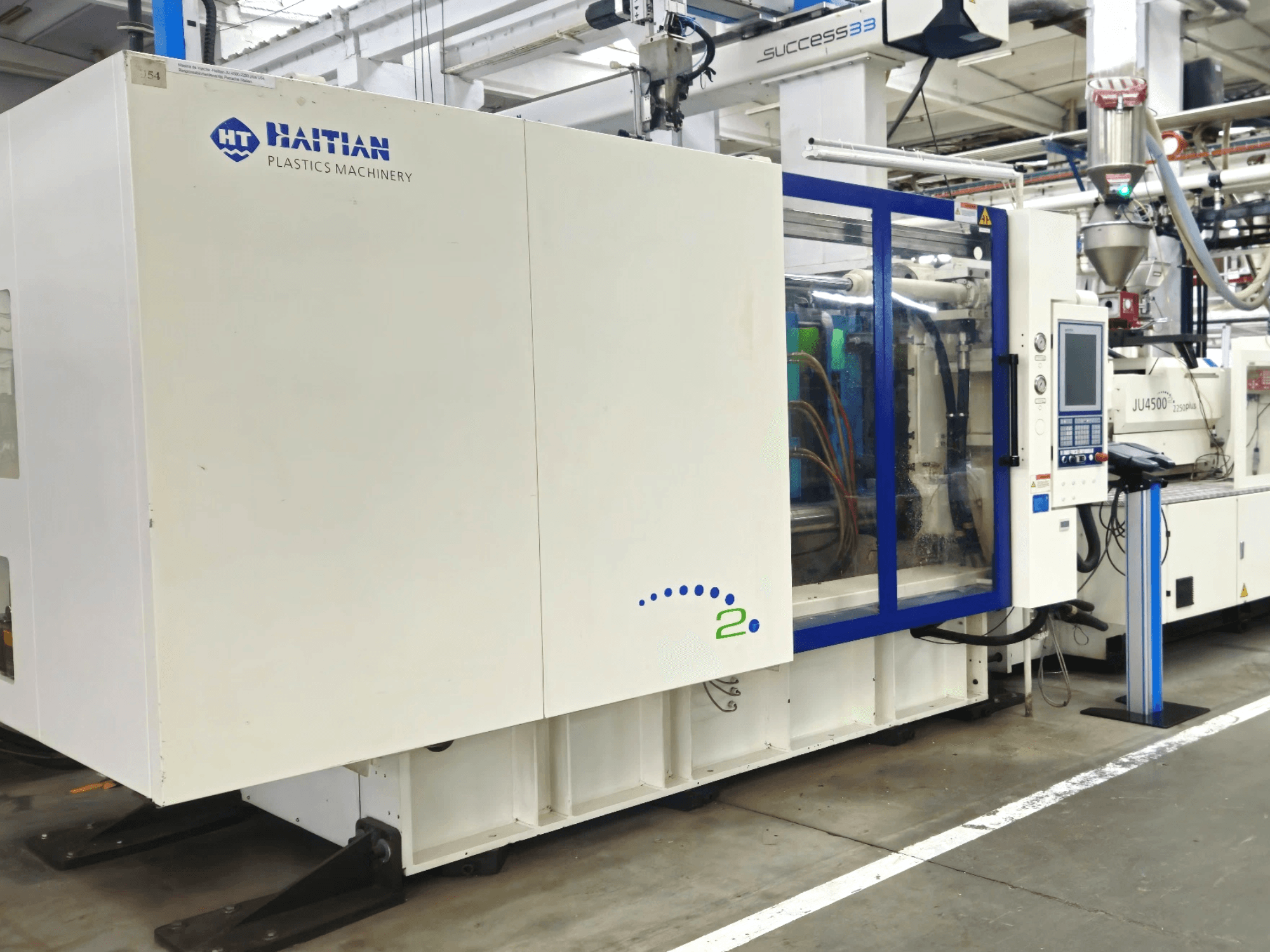 Haitian Plastics Machinery modell, framifrån, som visar maskiner med kontrollpanel och synliga slangar, i en industriell miljö.