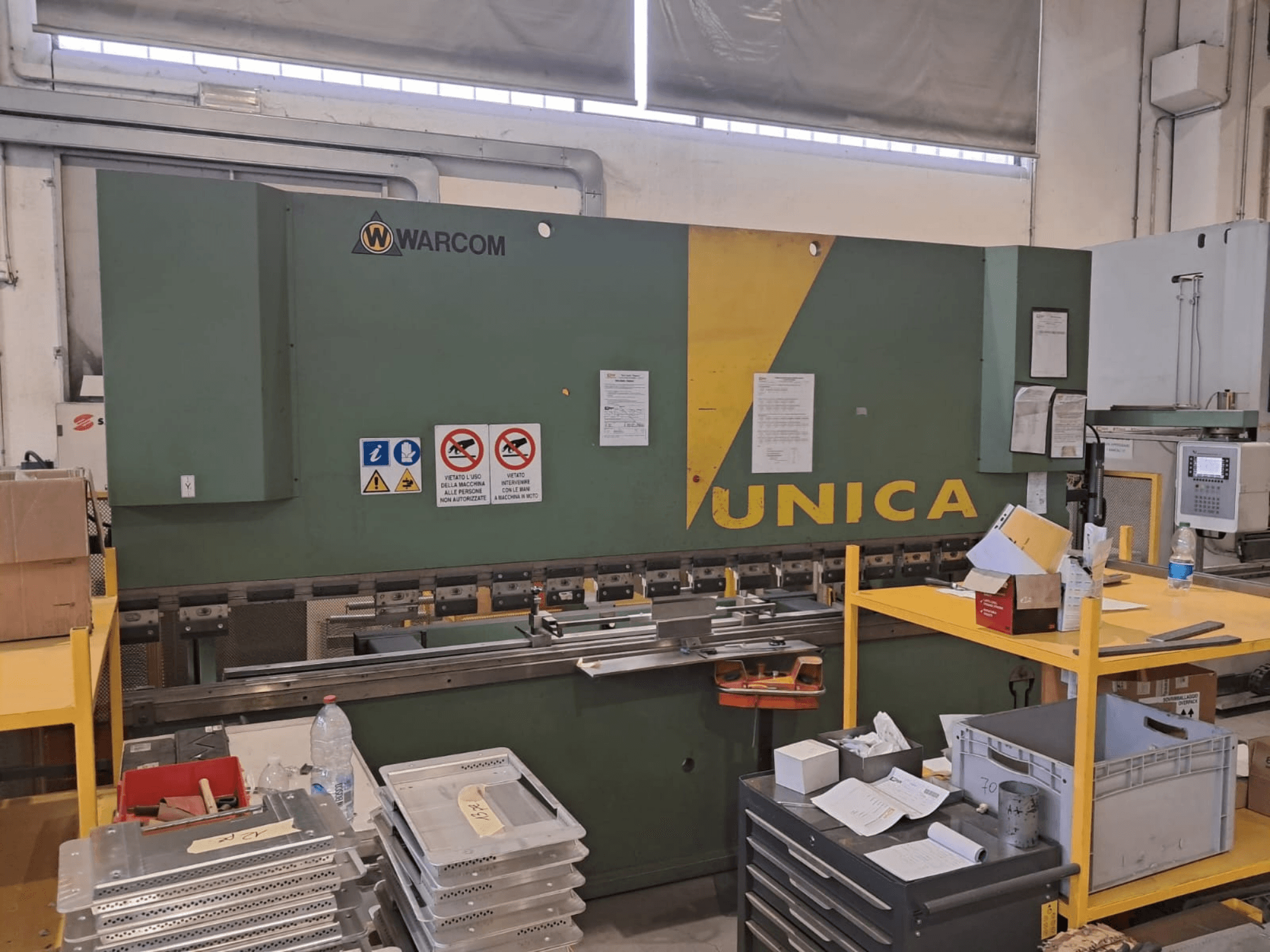 Hydraulisk press Warcom UNICA 100-40, framifrån, grön kaross med gula detaljer, säkerhetsskyltar synliga, verktyg och material på bordet.