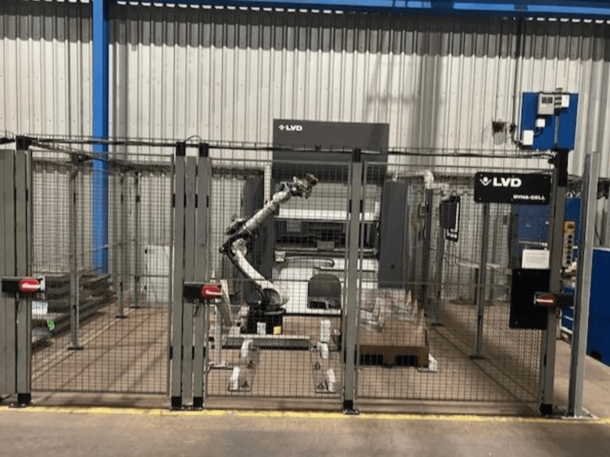 LVD Dyna-Cell robotarbetsstation i ett inhägnat område, framifrån, med robotarm och kontrollpanel.