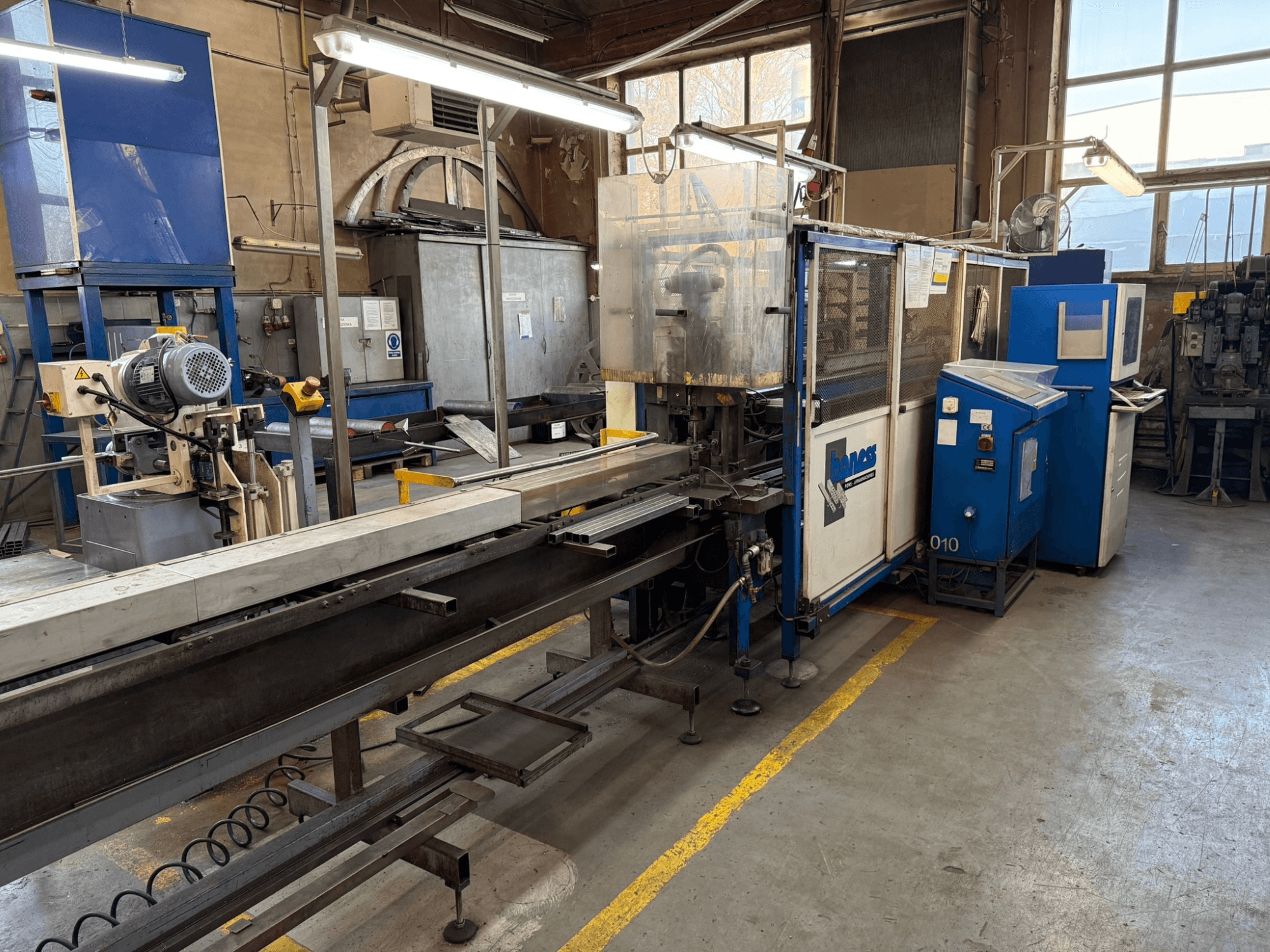 Industriverkstad med en blå Haweas-maskin, sidovy som visar transportband och utrustning i ett stort, väl upplyst utrymme.