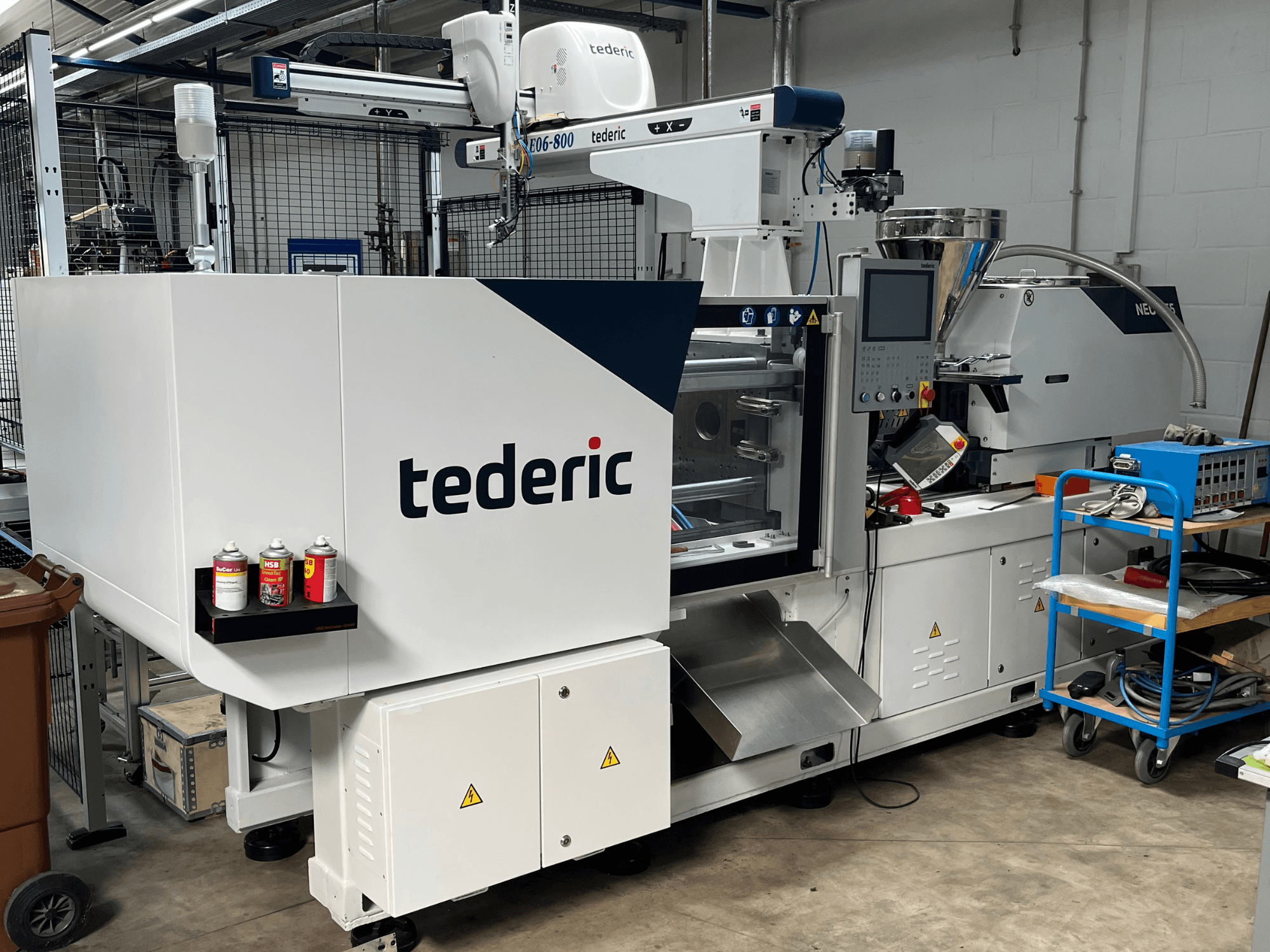Hydraulisk formsprutningsmaskin TEDERIC NEO.E55/e110h i en verkstad, sidovy med kontrollpanel och intilliggande verktyg.