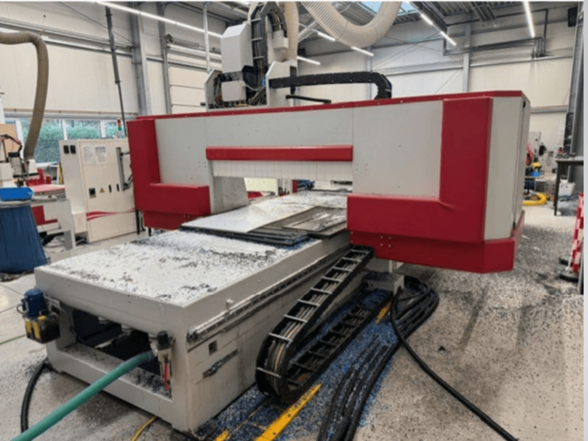 Röd och grå CNC-maskin i en verkstad, sidovy som visar materialbord, kylvätskeslang och sågspån på golvet.