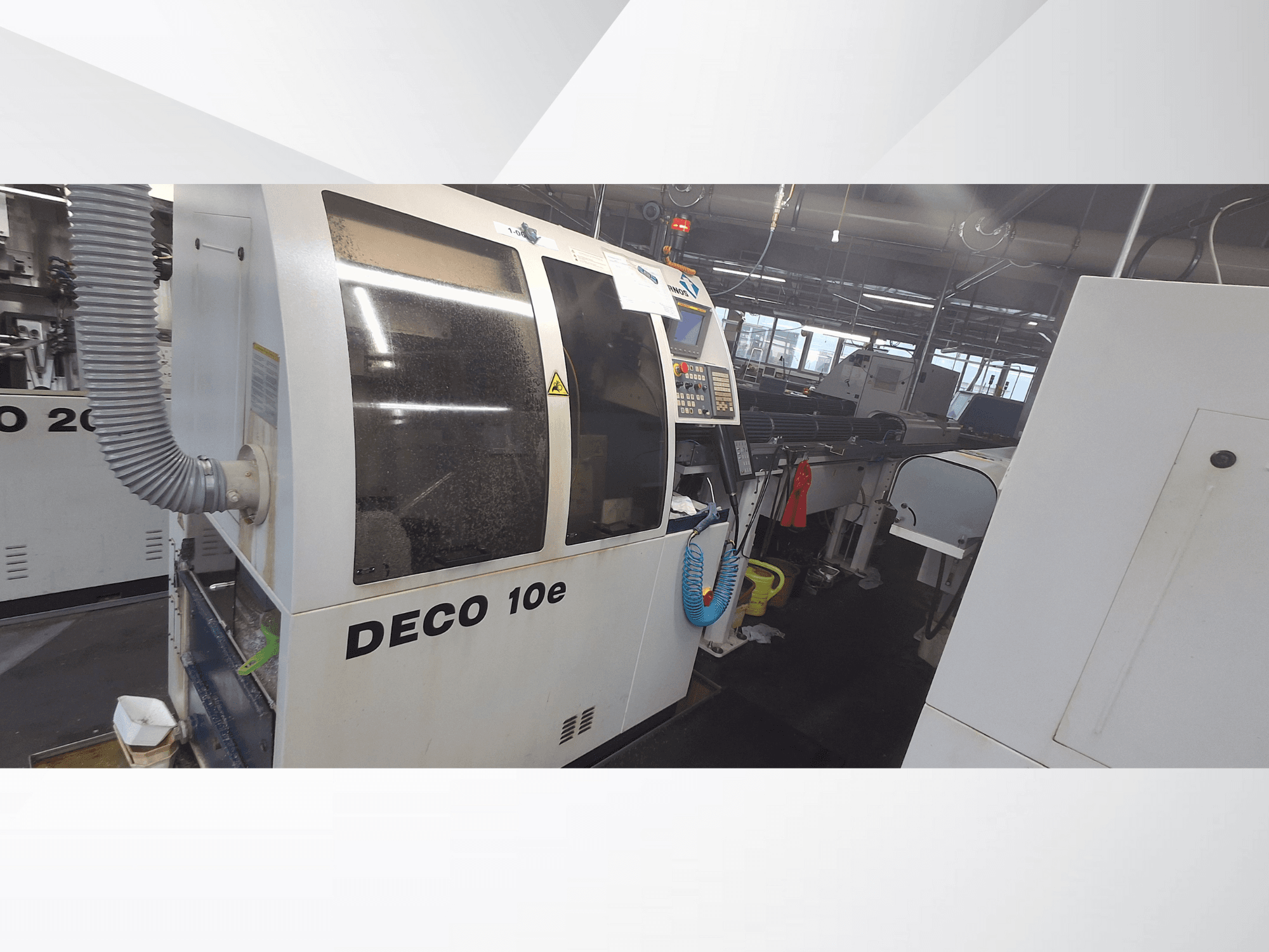 DECO 10e-maskin i en verkstad; sidovy som visar kontrollpanel och ventilationsslang, med suddiga maskiner i bakgrunden.