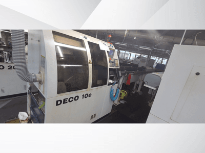 DECO 10e-maskin i en verkstad; sidovy som visar kontrollpanel och ventilationsslang, med suddiga maskiner i bakgrunden.