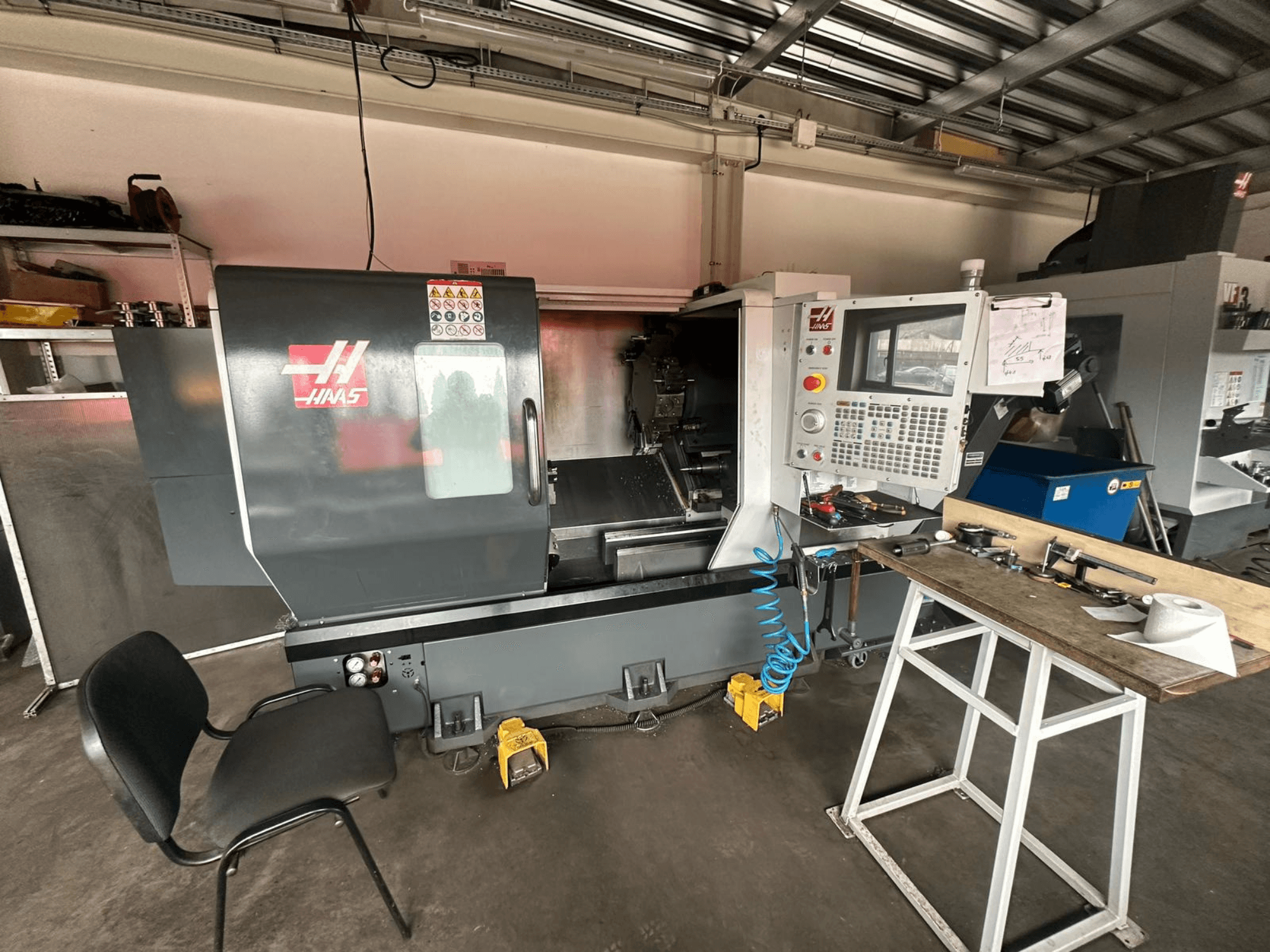 Haas CNC-svarv (modell ST-10) i en verkstad, sidovy som visar reglage, verktyg på en arbetsbänk och en stol i närheten.