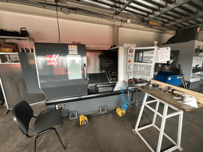 Haas CNC-svarv (modell ST-10) i en verkstad, sidovy som visar reglage, verktyg på en arbetsbänk och en stol i närheten.