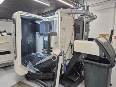 Öppen DMG-fräsmaskin med kylvätskespray, metallspån och verktygsreglage, sedd framifrån.
