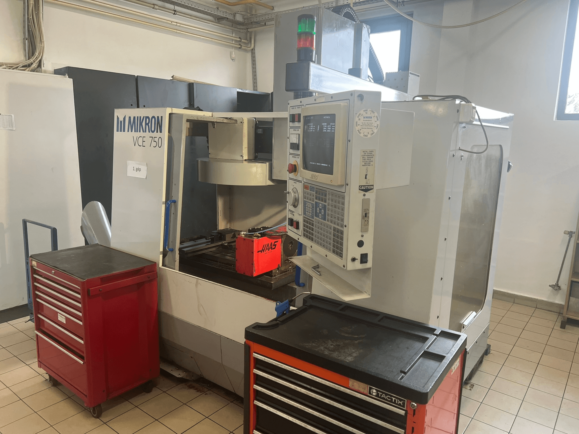 Alt text: "MIKRON VCE 750 Vertikal fleroperationsmaskin, framifrån med kontrollpanel och röda verktygslådor, i en rymlig verkstad."
