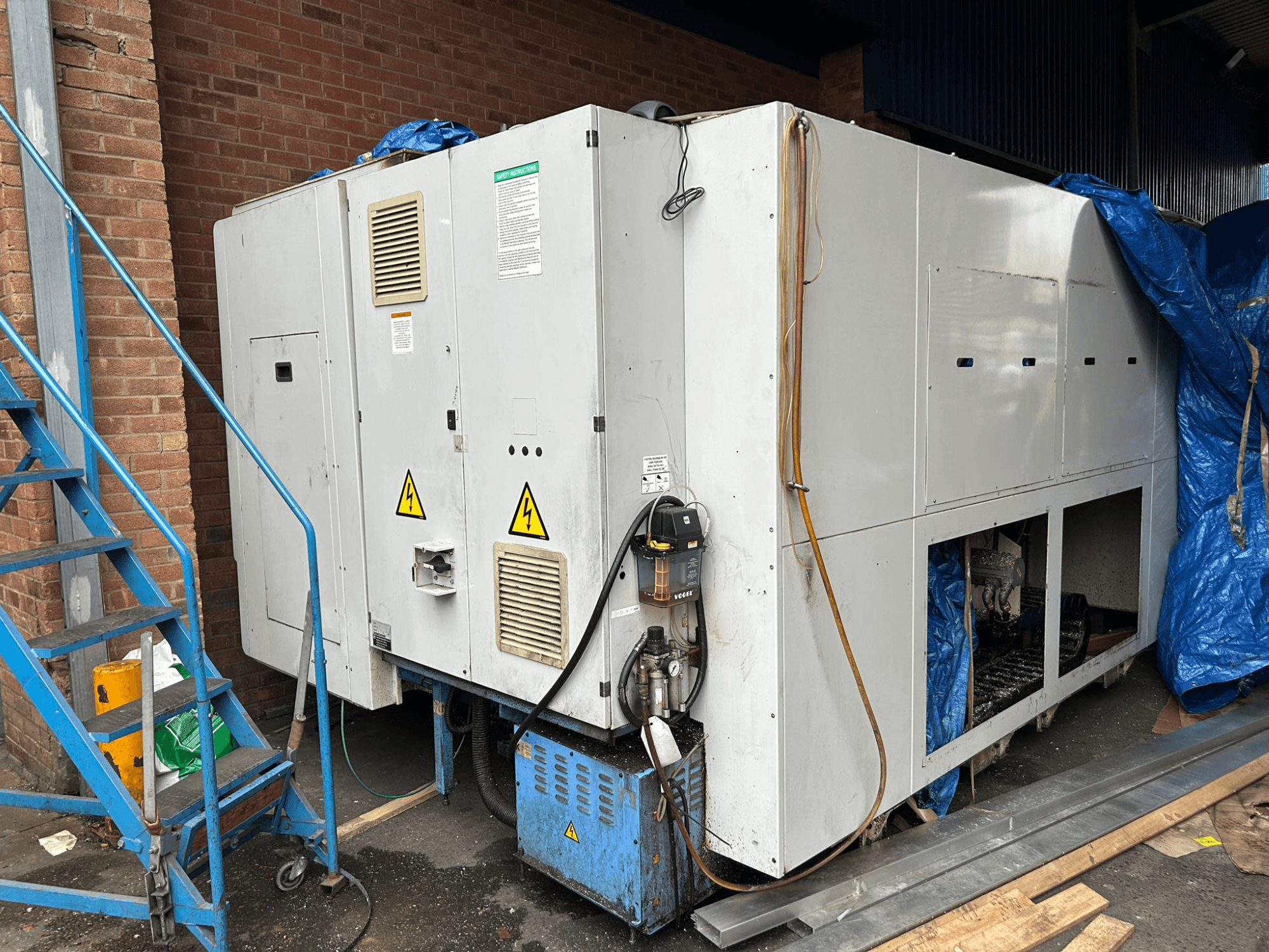 Sidovy av ett stort industriellt kylaggregat, märke och modell oklara, med elanslutningar, varningsskyltar och blå presenning.