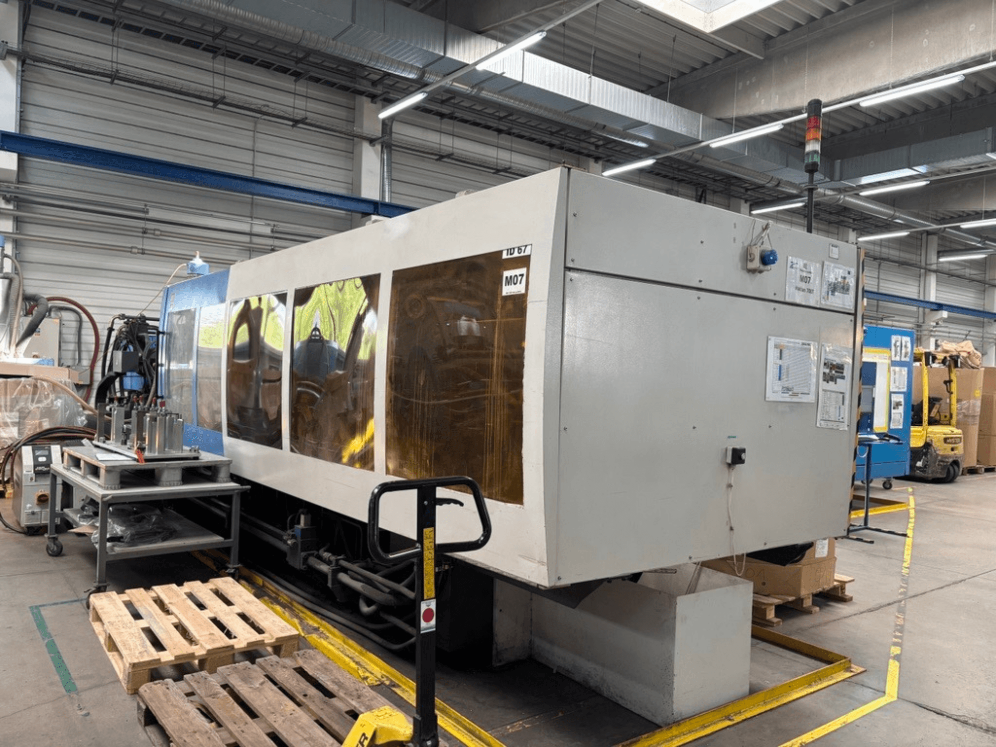 En hydraulisk formsprutningsmaskin av typen HAITIAN 7000/5000 i en fabrik, sett från sidan med kontrollpaneler och säkerhetslampor synliga.