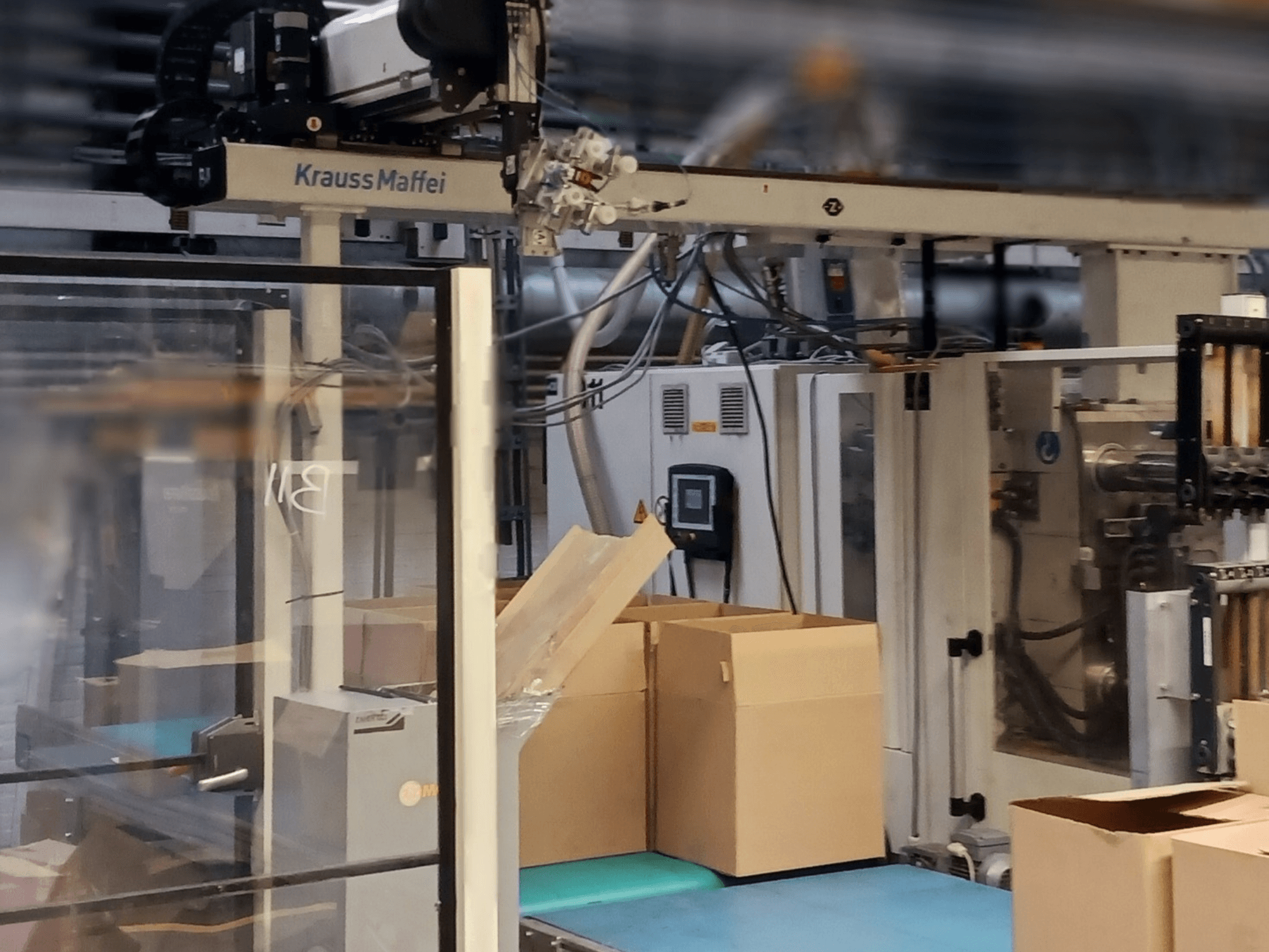 Krauss Maffei automationssystem i en fabrik, vy framifrån med lådor på en transportör och maskinkomponenter i drift.