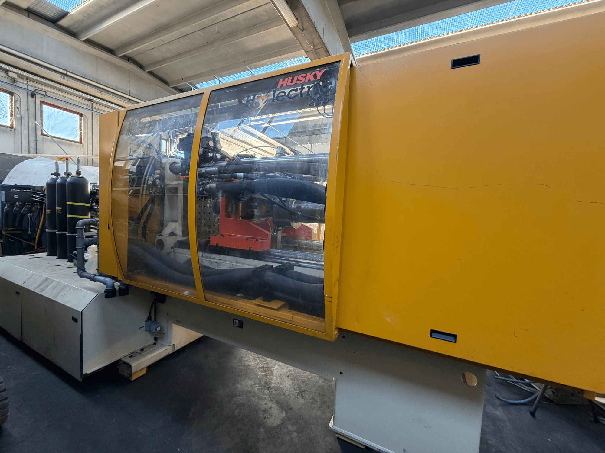 Husky HL225 RS65 elektrisk formsprutningsmaskin, framifrån med gult hölje, synlig kontrollpanel och hydrauliska komponenter.