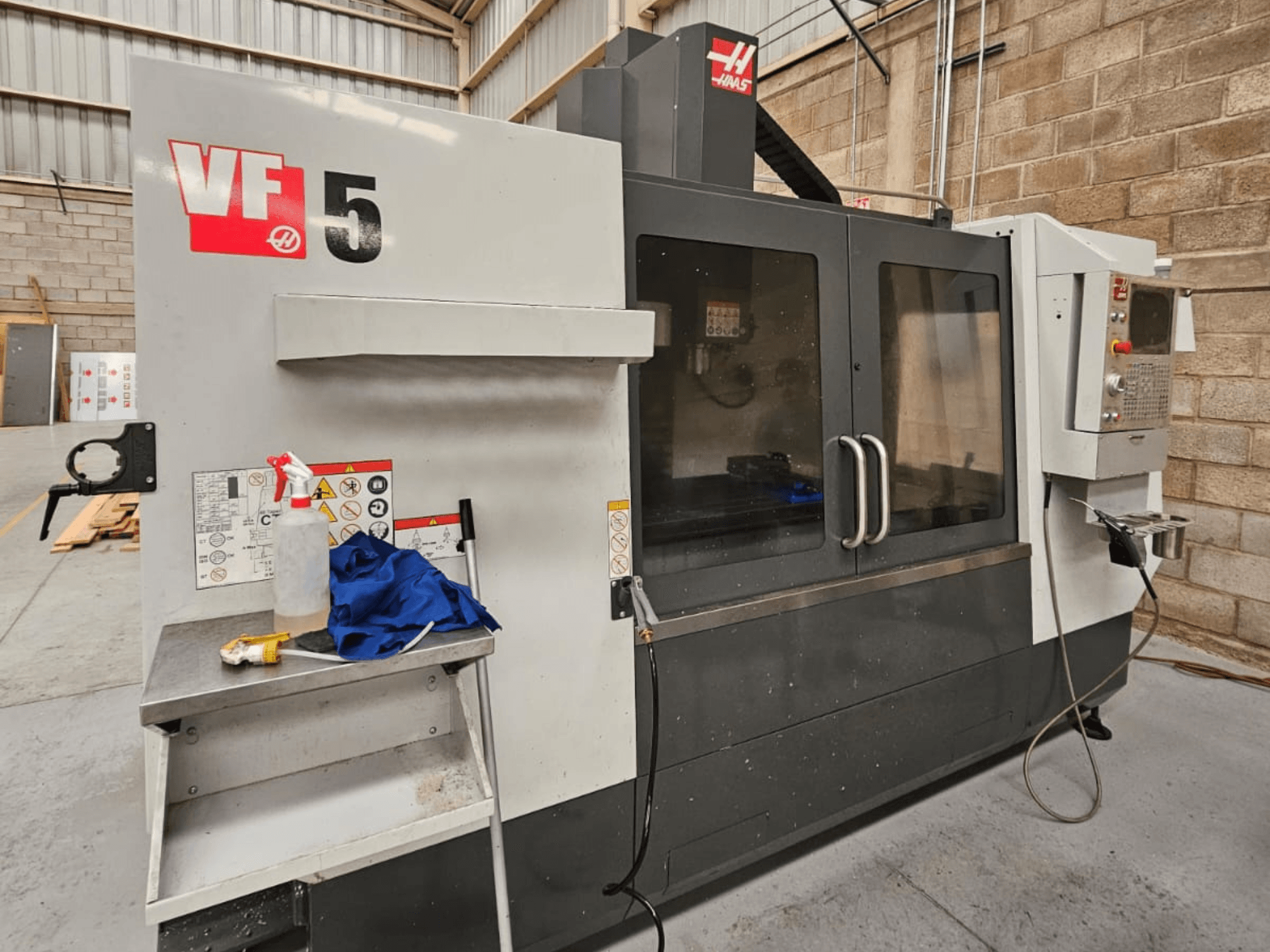 Haas VF-5 CNC-fräsmaskin framifrån, med kontrollpanel, kylvätskeflaska, blå trasa och säkerhetsinstruktioner.