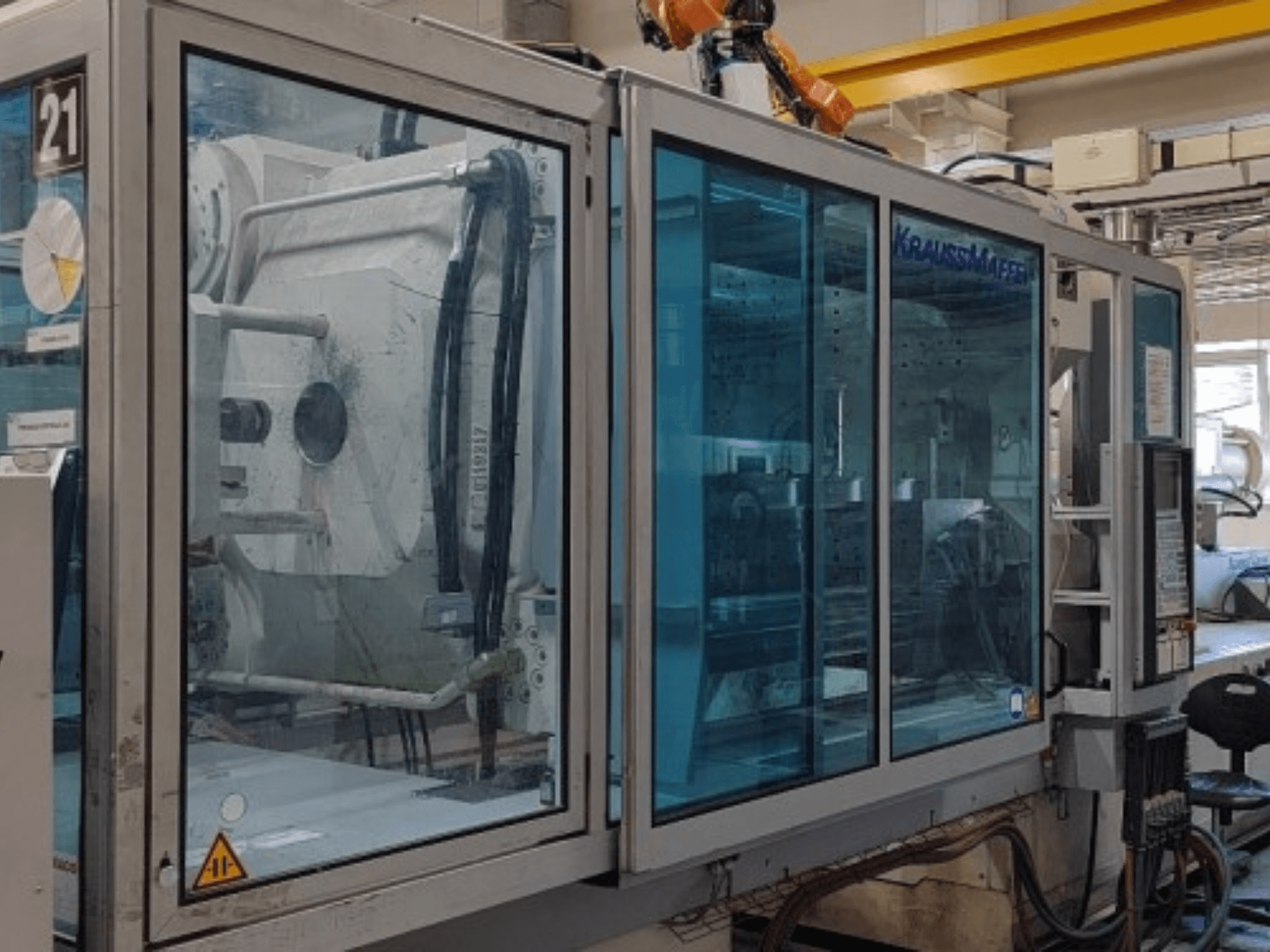 Hydraulisk formsprutningsmaskin av typen Krauss Maffei KM 575-2700 C2, sett från sidan, med genomskinliga säkerhetspaneler och manöverpanel.