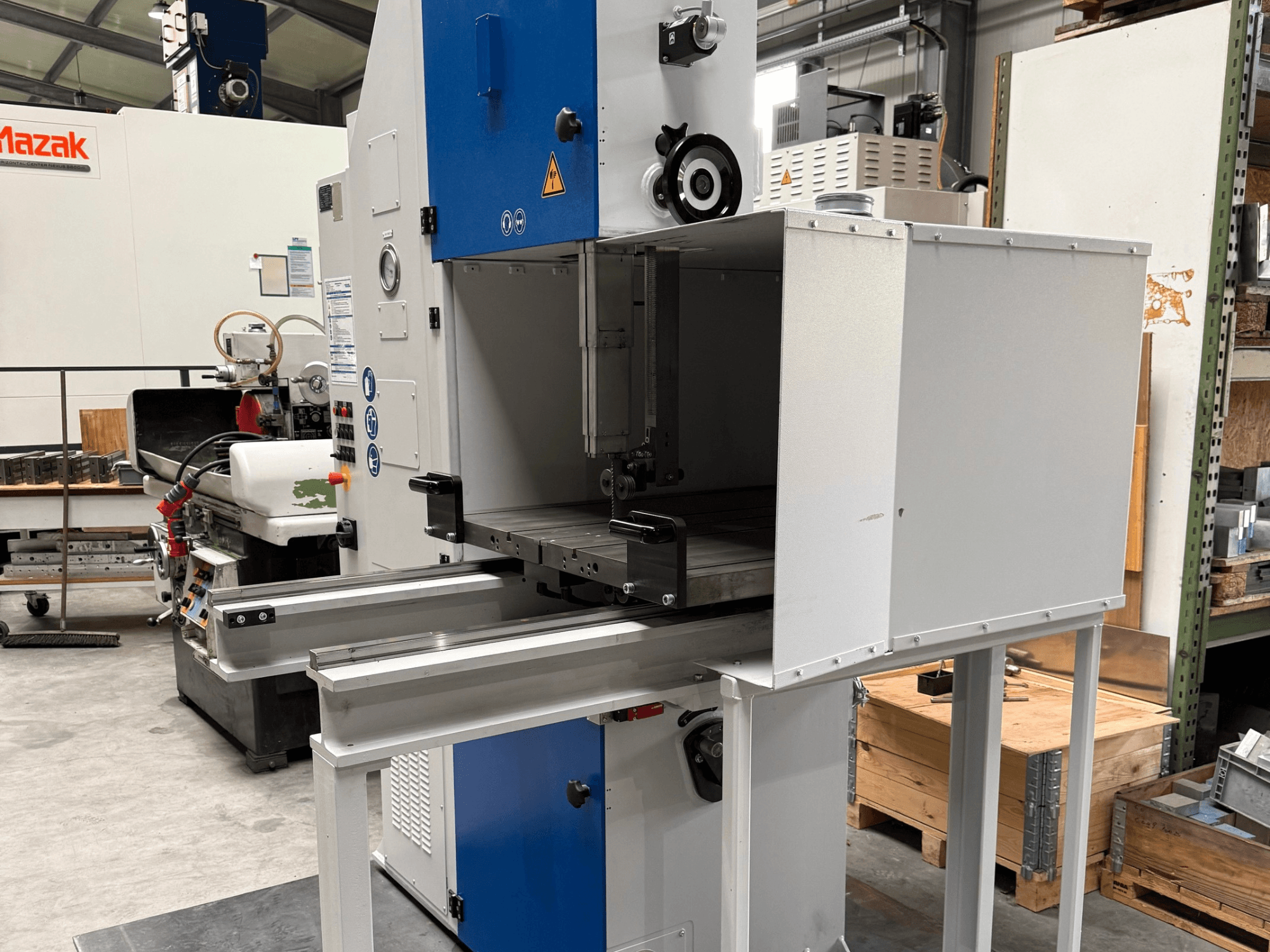 Mazak industrimaskin sedd framifrån, med blå kontrollpanel, metallbord och säkerhetsvarningar.