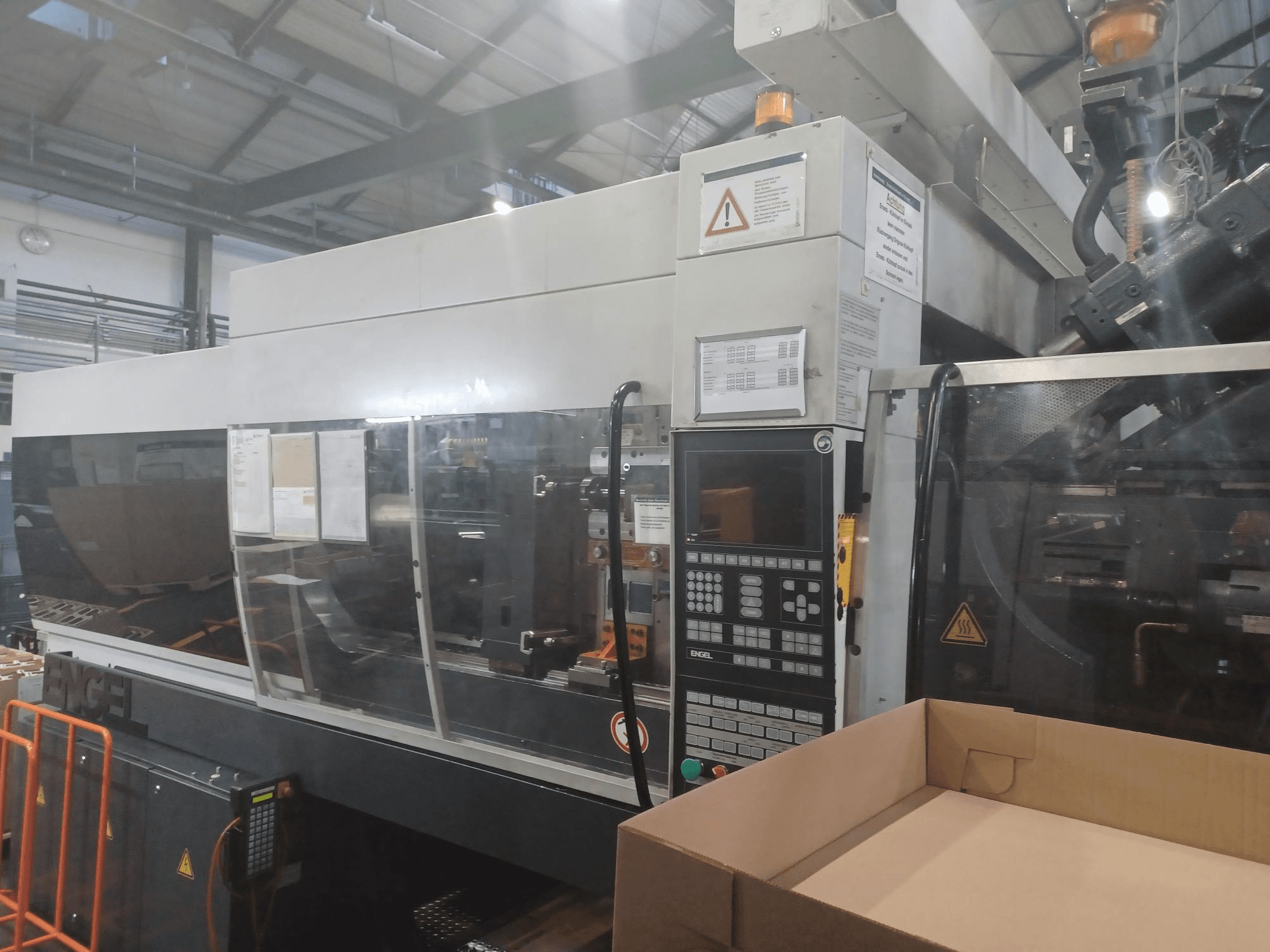 Engel formsprutningsmaskin, vy framifrån, med kontrollpanel, säkerhetsetiketter och en tom låda placerad i närheten.