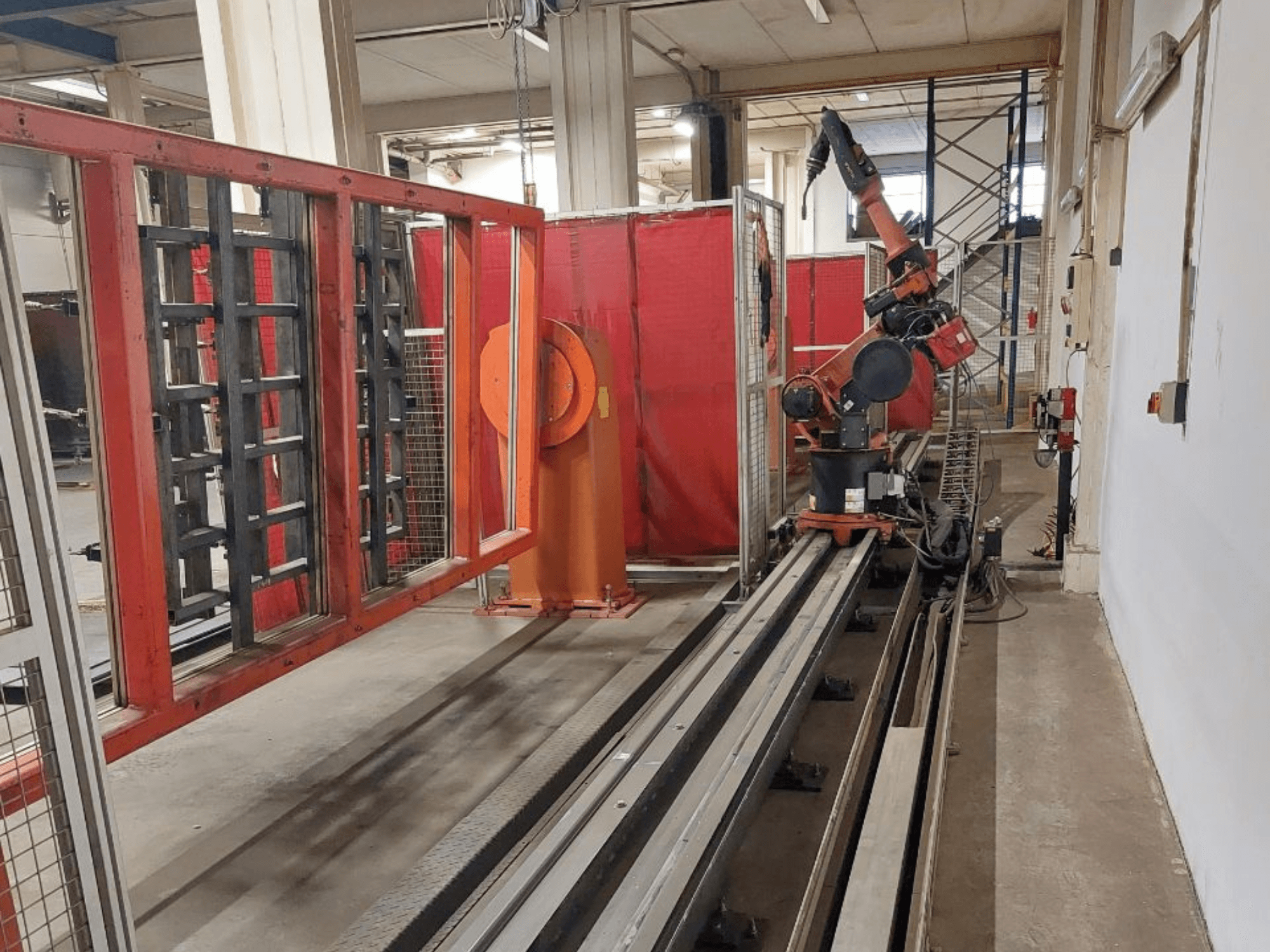 Industriell robotarm med orange och svarta färger placerad på metallskenor, omgiven av röda skyddsbarriärer, vy framifrån.