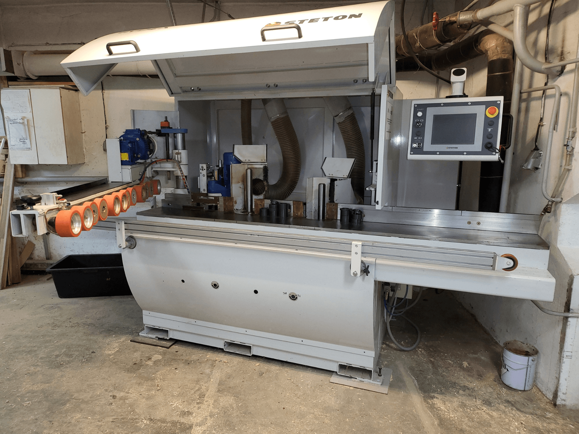 En frontvy av en Stetton träbearbetningsmaskin med kontrollpanel, matarrör och olika trästycken på bordet.