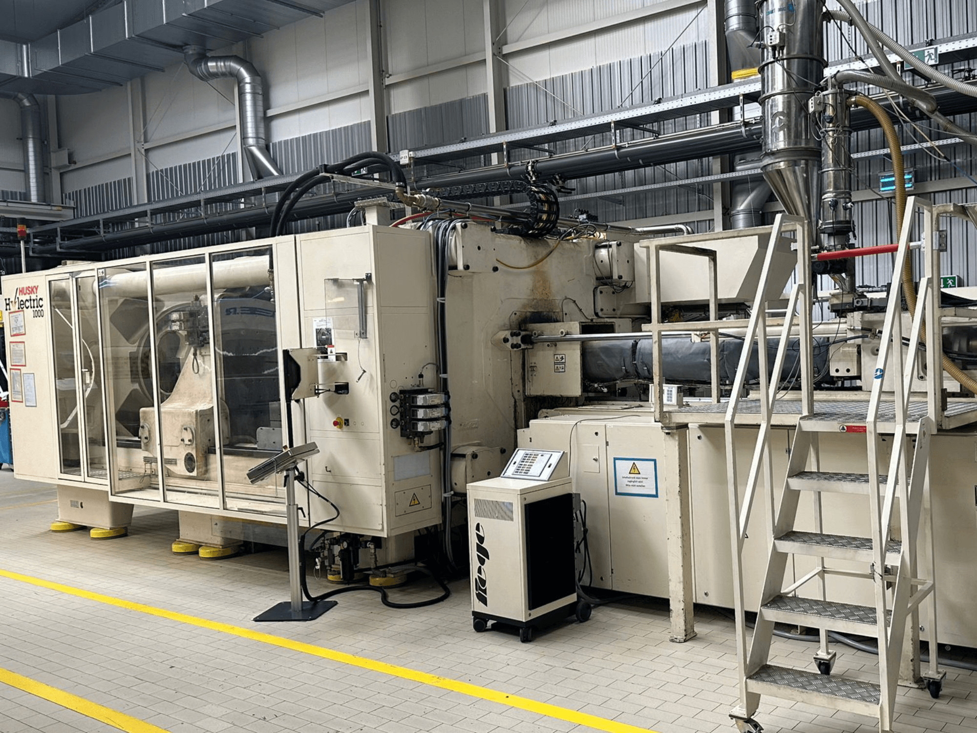 Framifrånvy av den elektriska formsprutningsmaskinen Husky Hylelectric H1000 RS95/80 i en fabriksmiljö, med kontrollpanel och trappsteg.