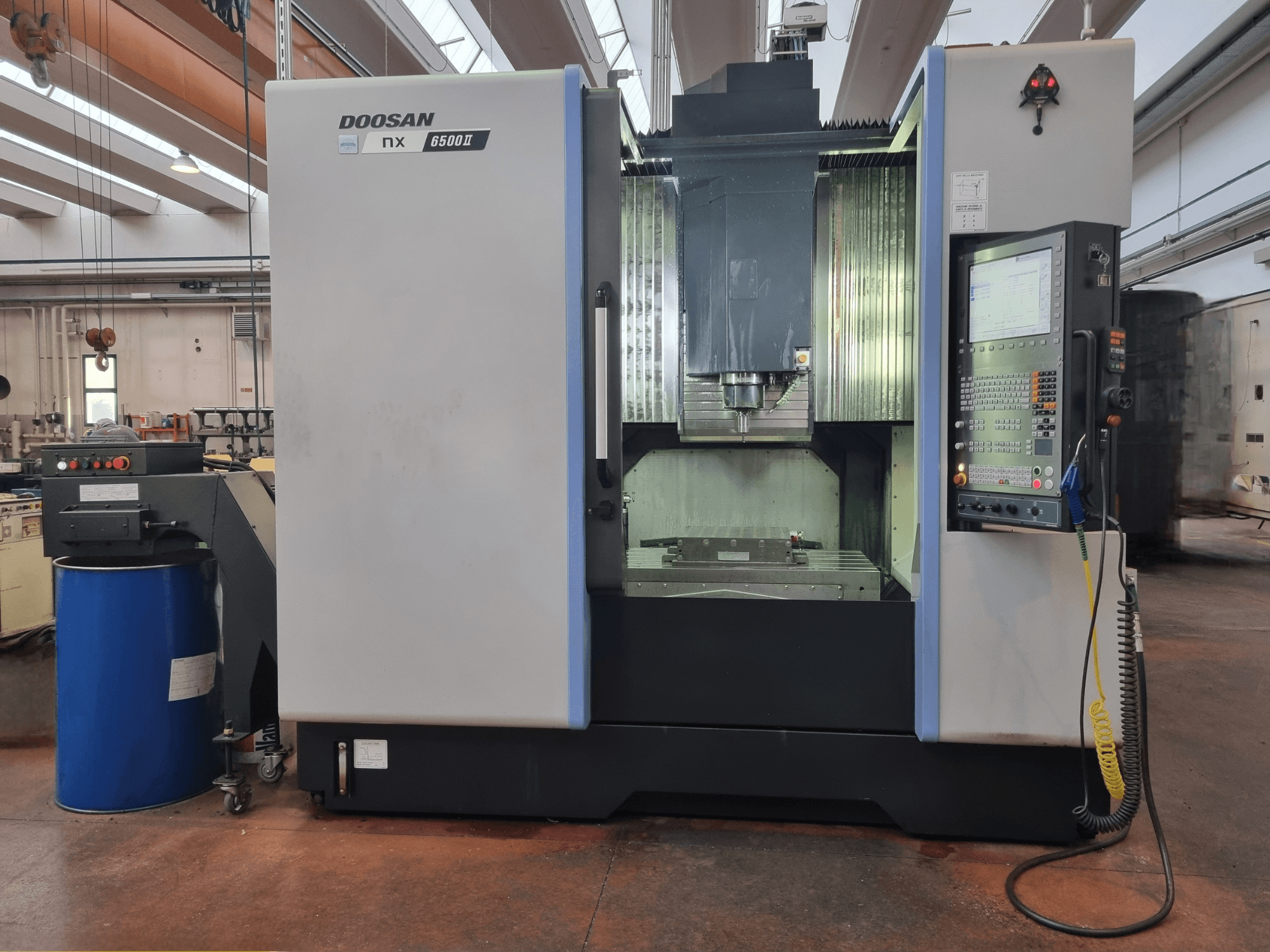 Doosan NX 6500 II CNC-maskin, framifrån, med kontrollpanel och bearbetningsområde, placerad bredvid en blå tunna.
