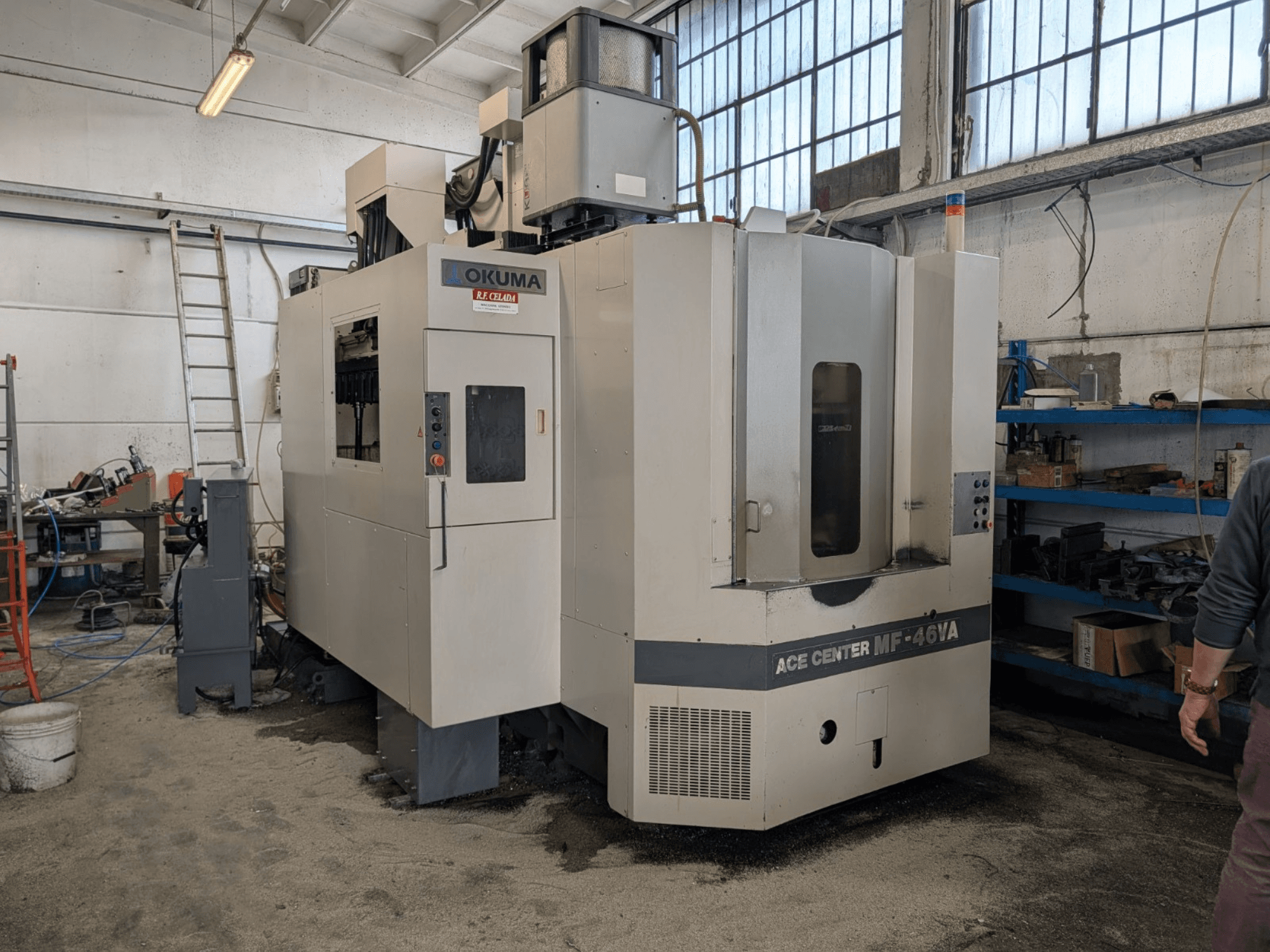 Okuma ACE CENTER MF-46VA vertikal fleroperationsmaskin i en verkstad, framifrån med kontrollpanel och omgivande utrustning.