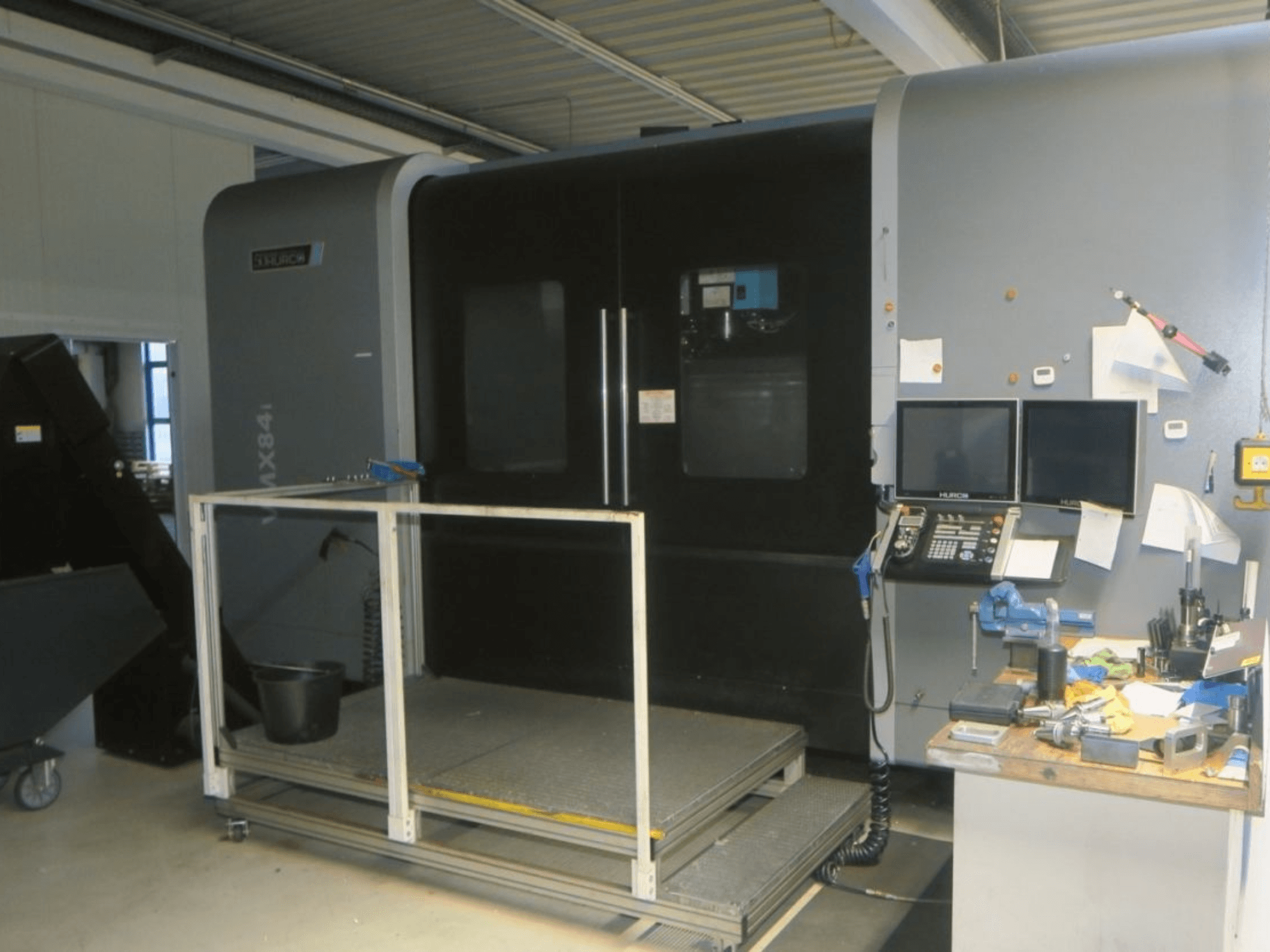 Hurco MX84 CNC-maskin framifrån, med dubbla bildskärmar, kontrollpanel och verktygsarbetsyta med olika verktyg.