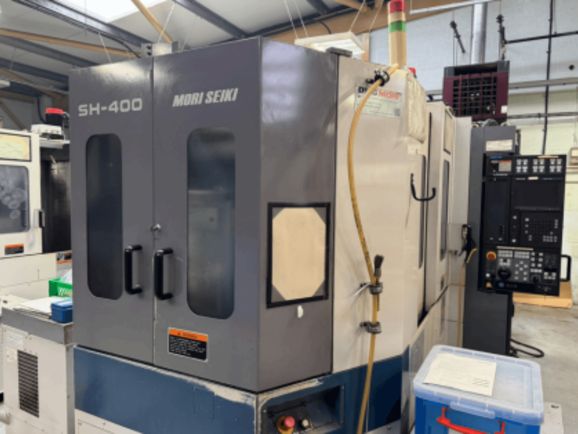 Mori Seiki SH-400 CNC-maskin, framifrån, med dubbla dörrar, kontrollpanel och tillbehörsslang. Blå förvaringsbehållare i närheten.