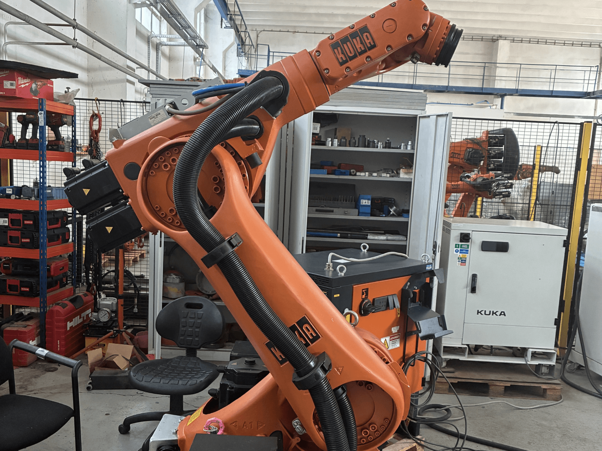 KUKA KR30-3 robotarm i en industriell miljö, sidovy som visar kablar och reglage, med verktyg och hyllor i bakgrunden.