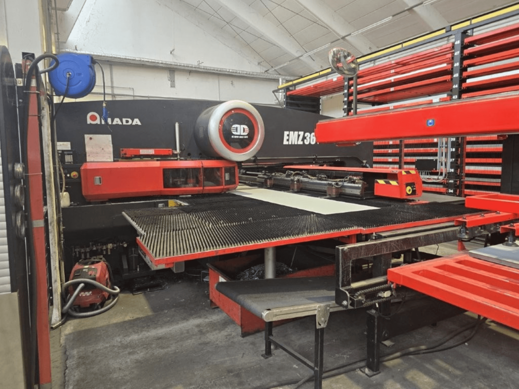 Amada EMZ 36 laserskärmaskin i en verkstad, framifrån, med röda detaljer och ett stort skärbord.